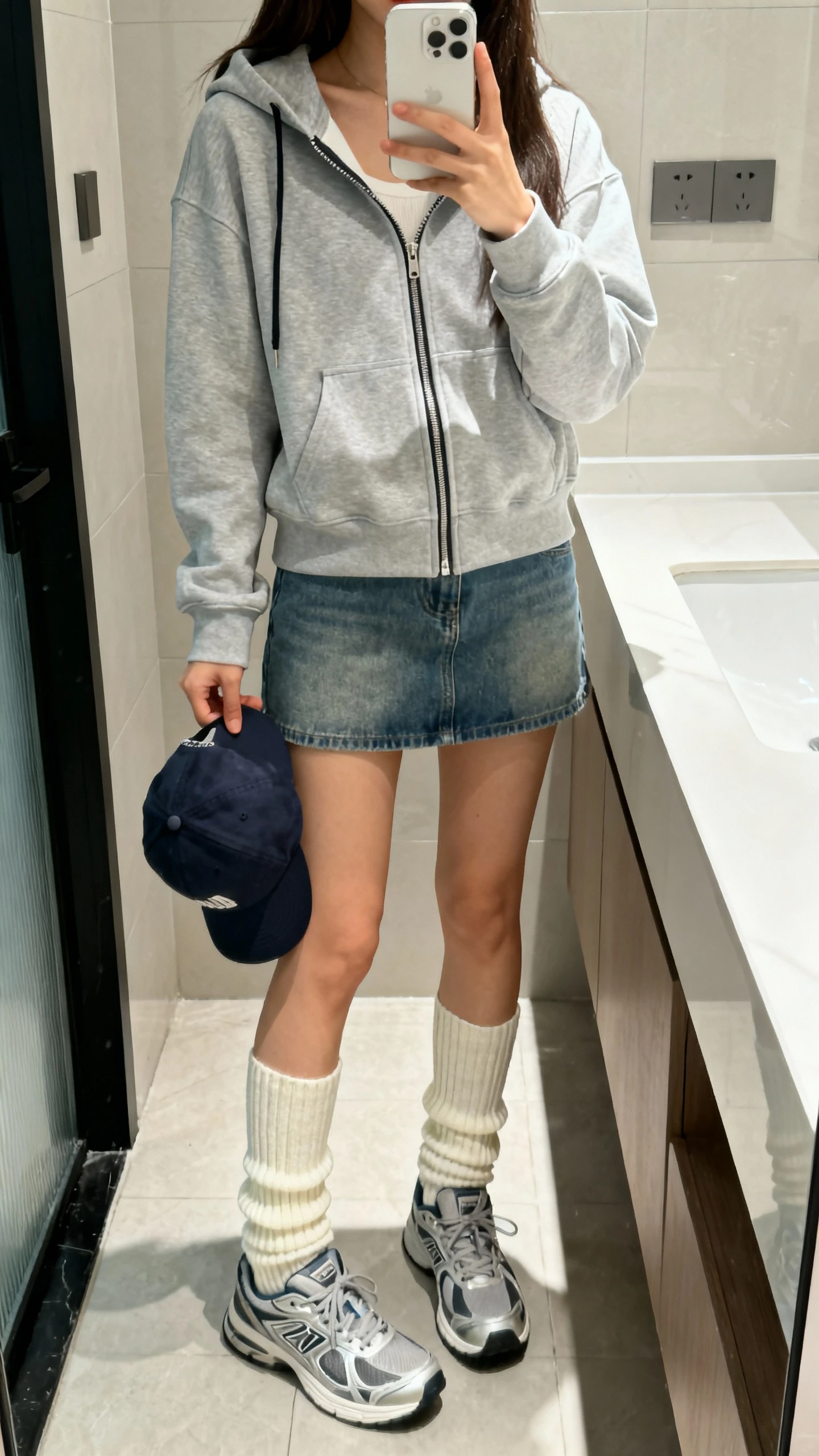 iPhone mirror selfie of a woman wearing a zip-up hoodie over a denim mini skirt with high crew socks and sporty sneakers, baseball cap in hand, face not visible, bathroom mirror, bright indoor lighting, iPhone photo quality.