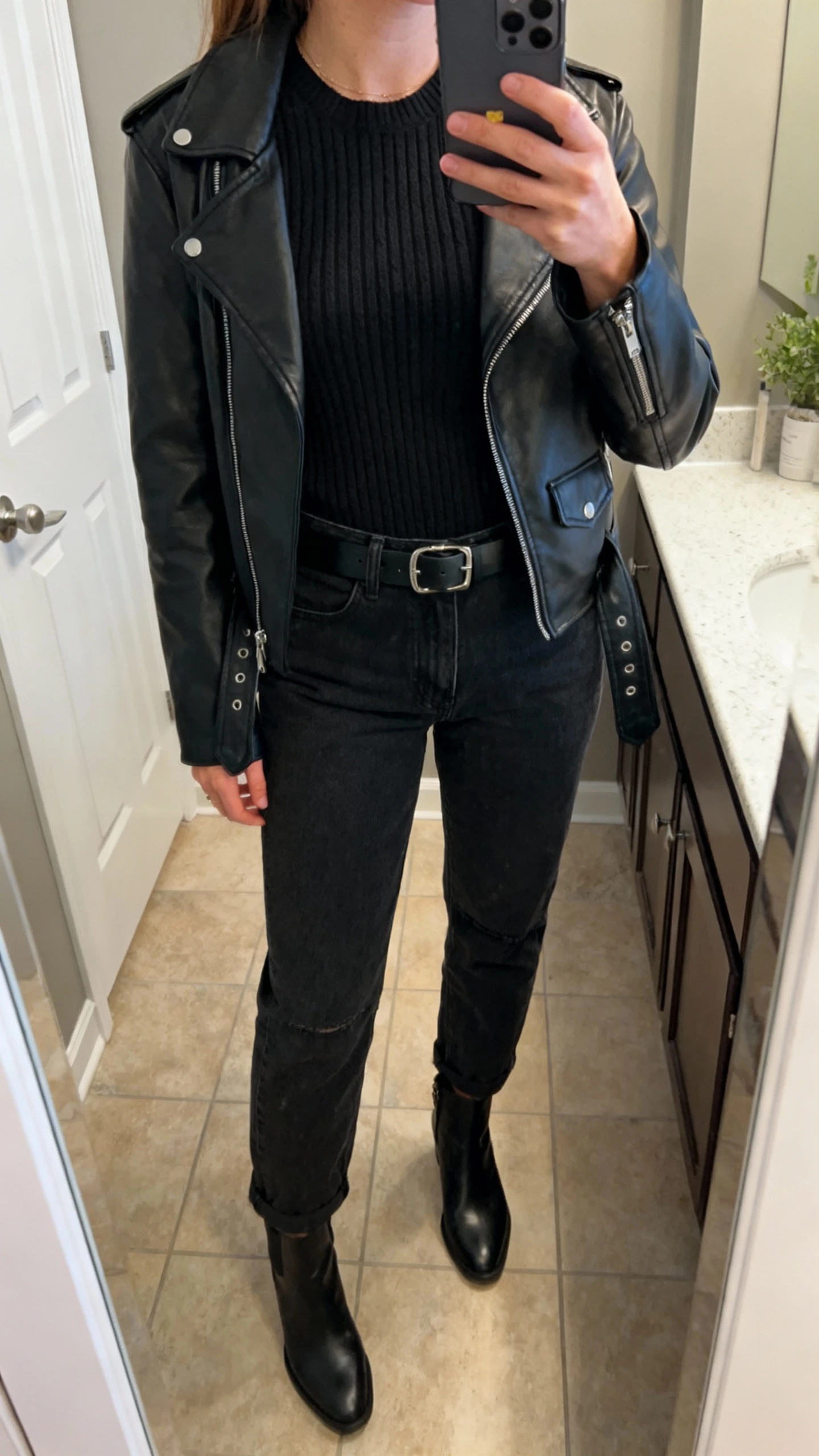 iPhone mirror selfie of a woman wearing all-black: black mom jeans, a black textured knit or ribbed top, a black leather moto jacket, black ankle boots, and a black belt, face not visible, cropped at neck in bathroom mirror, indoor lighting, iPhone photo quality.