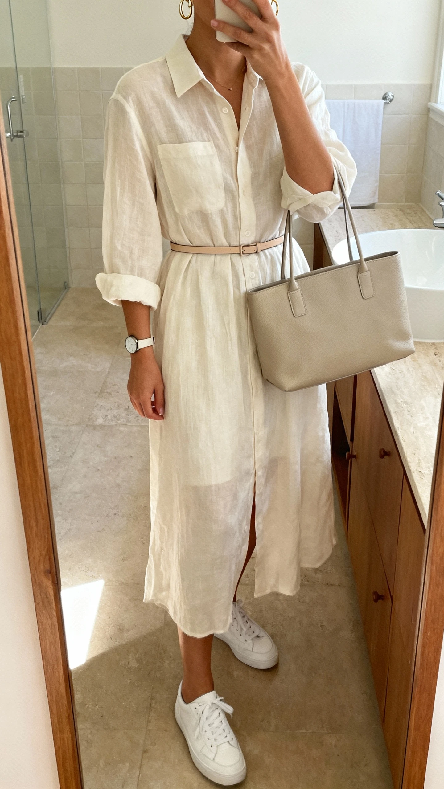 iPhone mirror selfie of a woman wearing an airy shirt dress cinched with a slim belt, simple white sneakers, structured tote, delicate hoop earrings, and a minimalist watch, face not visible, cropped at shoulders, bathroom mirror, soft indoor lighting, iPhone photo quality