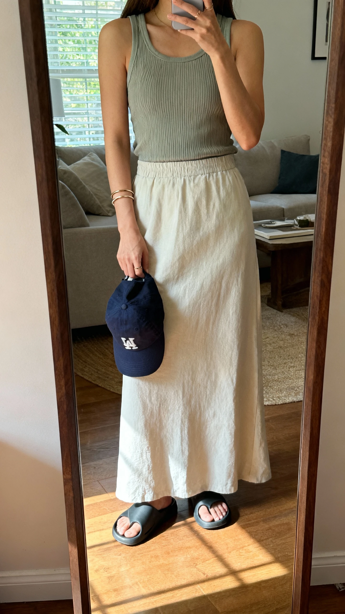 iPhone mirror selfie of a woman wearing an effortless maxi skirt with a sporty ribbed tank, rubber slides, baseball cap tucked in hand, simple bangles, face not visible, full-length mirror in living room, soft afternoon window light, iPhone photo quality.