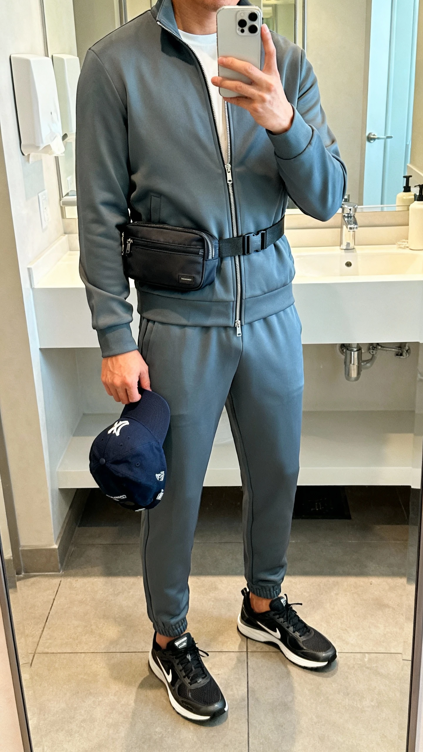 iPhone mirror selfie of a woman wearing an elevated athleisure set (tailored zip-up track jacket and matching tapered joggers), sleek trainers, crossbody belt bag, baseball cap held in hand, face not visible, bathroom mirror, soft indoor lighting, iPhone photo quality.