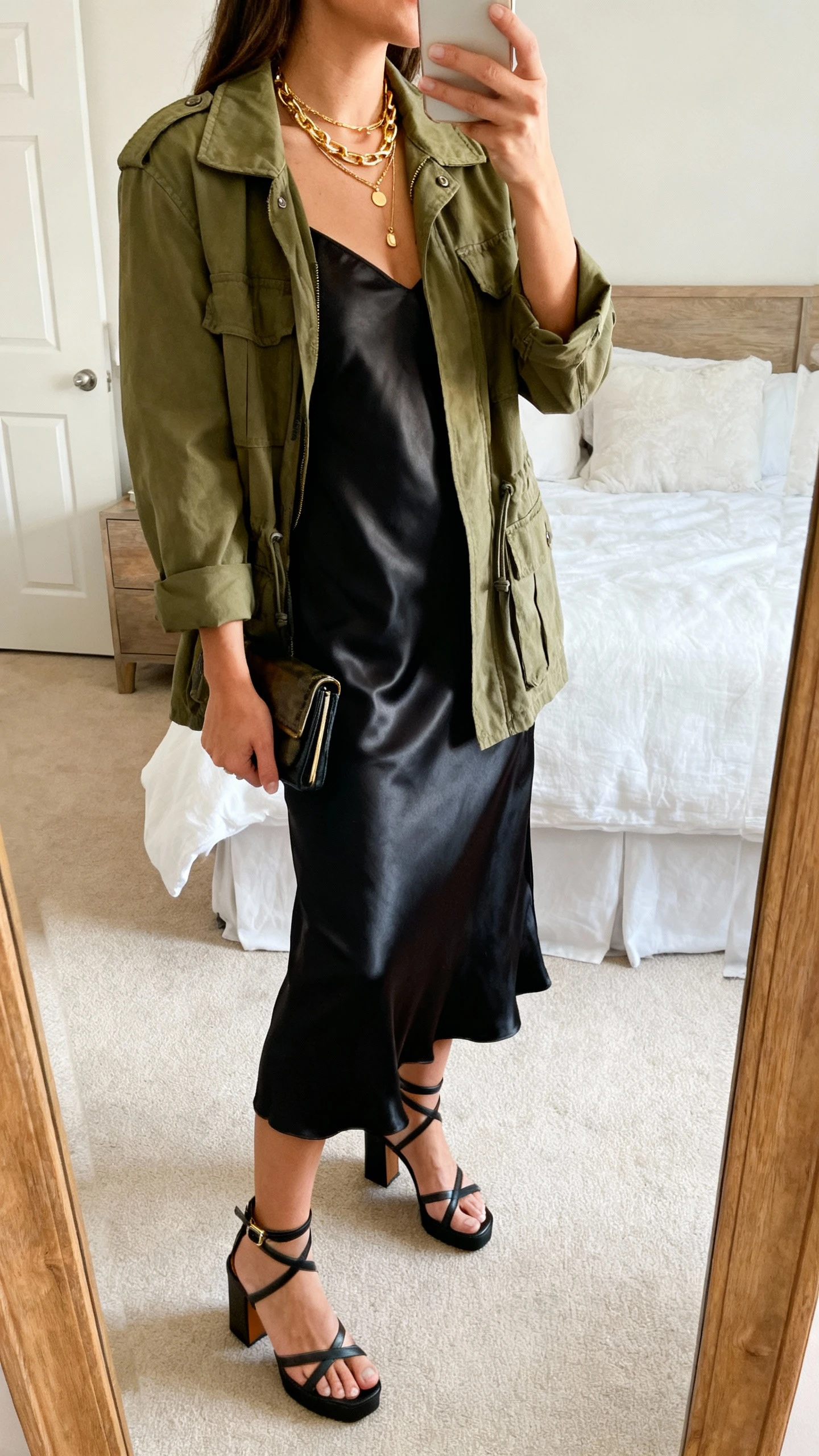iPhone mirror selfie of a woman wearing an olive utility jacket over a satin black slip dress, strappy block-heel sandals, layered gold necklaces, and a small clutch, face not visible, side angle, full-length mirror in bedroom, natural morning light, iPhone photo quality.