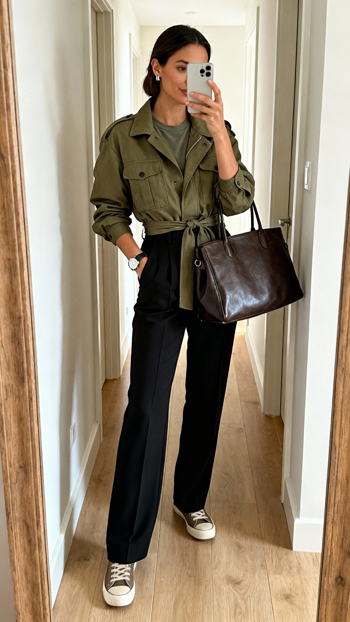 iPhone mirror selfie of a woman wearing an olive utility jacket with cinched waist, high-waisted black tailored trousers, minimal leather sneakers, simple crewneck tee, structured leather tote, sleek stud earrings, and a watch; face not visible, hallway mirror, natural morning light, iPhone photo quality.