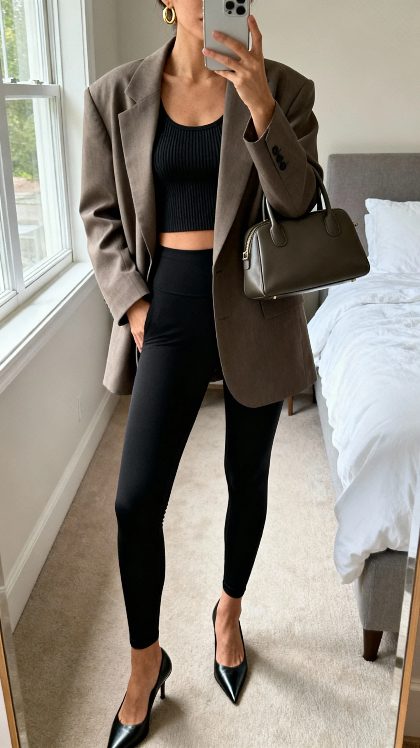 iPhone mirror selfie of a woman wearing an oversized tailored blazer over sleek black high-waisted leggings, fitted ribbed tank, pointed-toe heels, structured tote, minimal gold hoops, face not visible, cropped at neck, bedroom mirror, natural window light, iPhone photo quality.