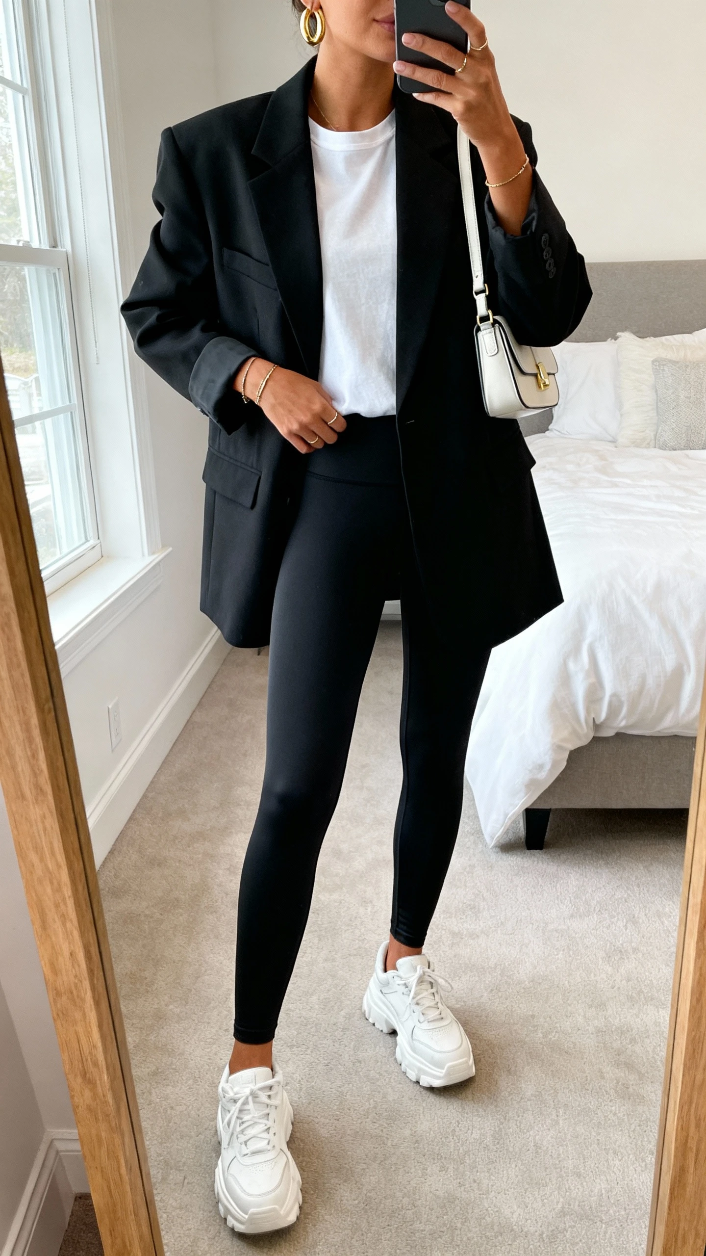 iPhone mirror selfie of a woman wearing an oversized structured blazer in black over a basic white crewneck tee, high-waisted matte black leggings, chunky white sneakers, a mini shoulder bag, gold hoop earrings and minimal jewelry, face not visible, cropped at neck, bedroom mirror, natural window light, iPhone photo quality.