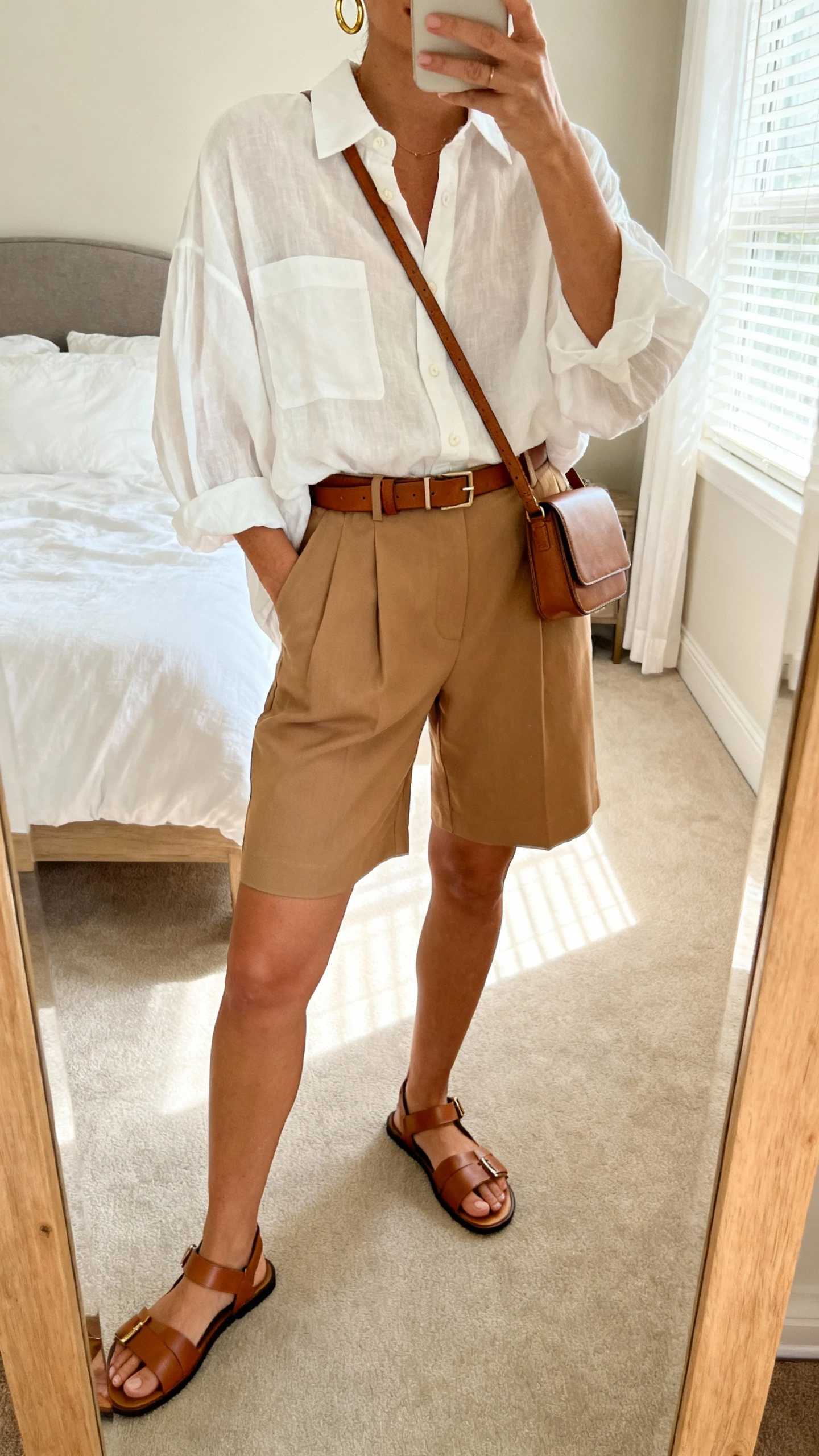 iPhone mirror selfie of a woman wearing an oversized breezy white button-up tucked into tailored tan shorts, slim leather belt, minimalist gold hoops, structured crossbody, and sleek leather sandals, face not visible, cropped at neck, bedroom mirror, morning window light, iPhone photo quality.