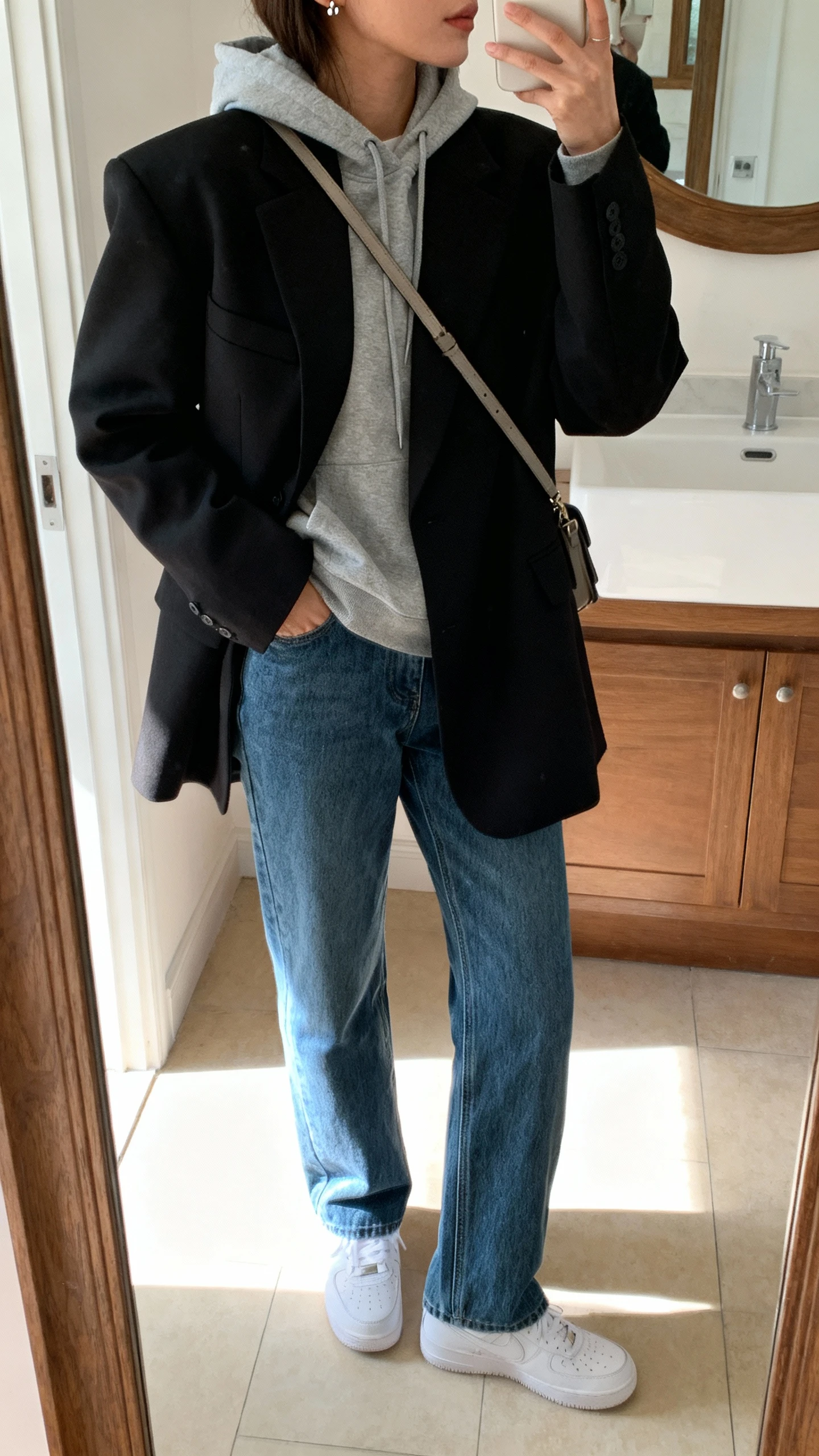 iPhone mirror selfie of a woman wearing an oversized black blazer over a gray hoodie, straight blue jeans, white sneakers, crossbody bag, and simple studs, face not visible, side angle crop at shoulders, bathroom mirror, indoor morning light, iPhone photo quality.