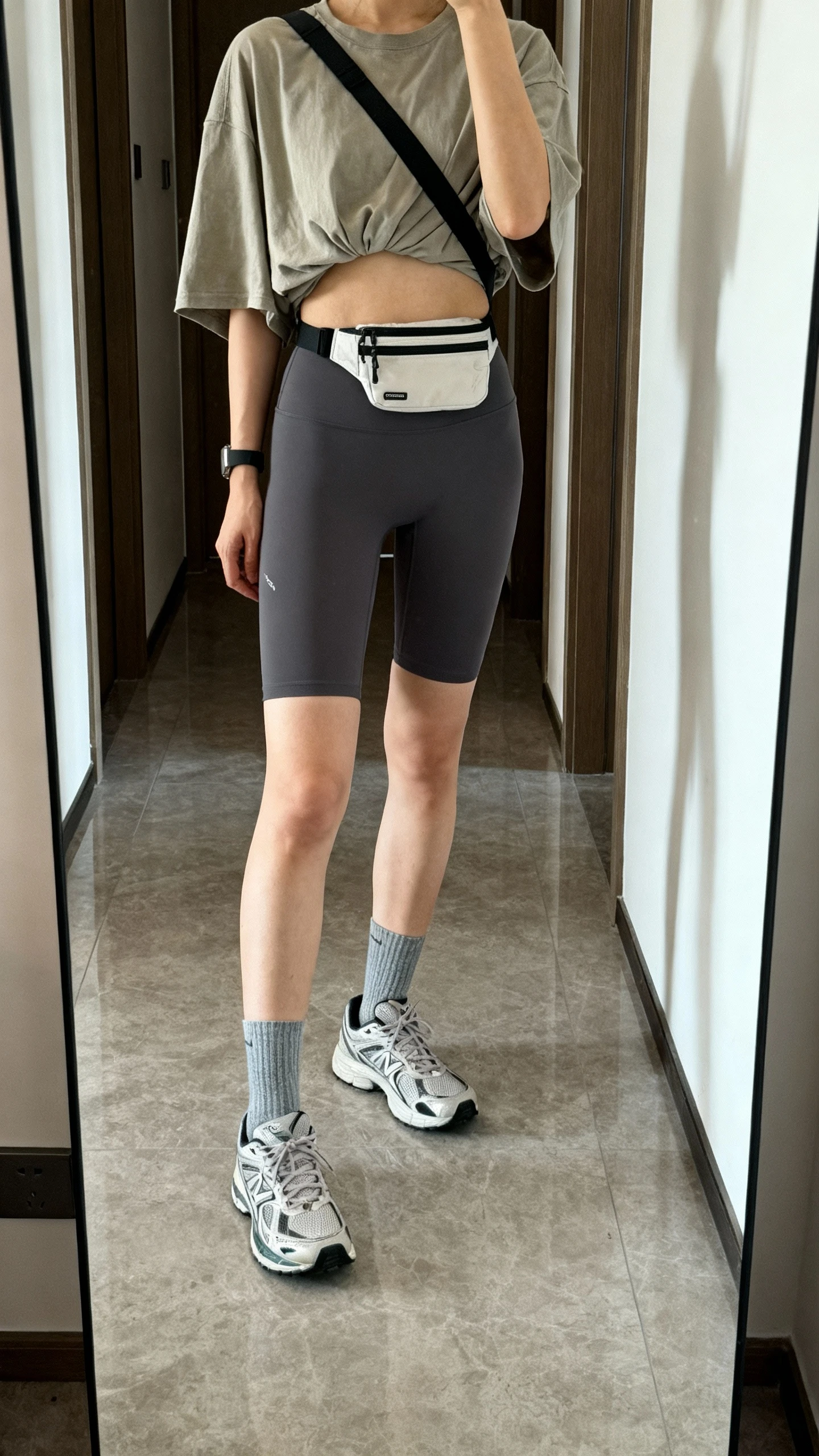 iPhone mirror selfie of a woman wearing an oversize breezy tee half-tucked into compressive biker shorts, ankle socks, and running sneakers, small crossbody belt bag slung across, face not visible, cropped at shoulders, hallway mirror, natural daylight, iPhone photo quality.