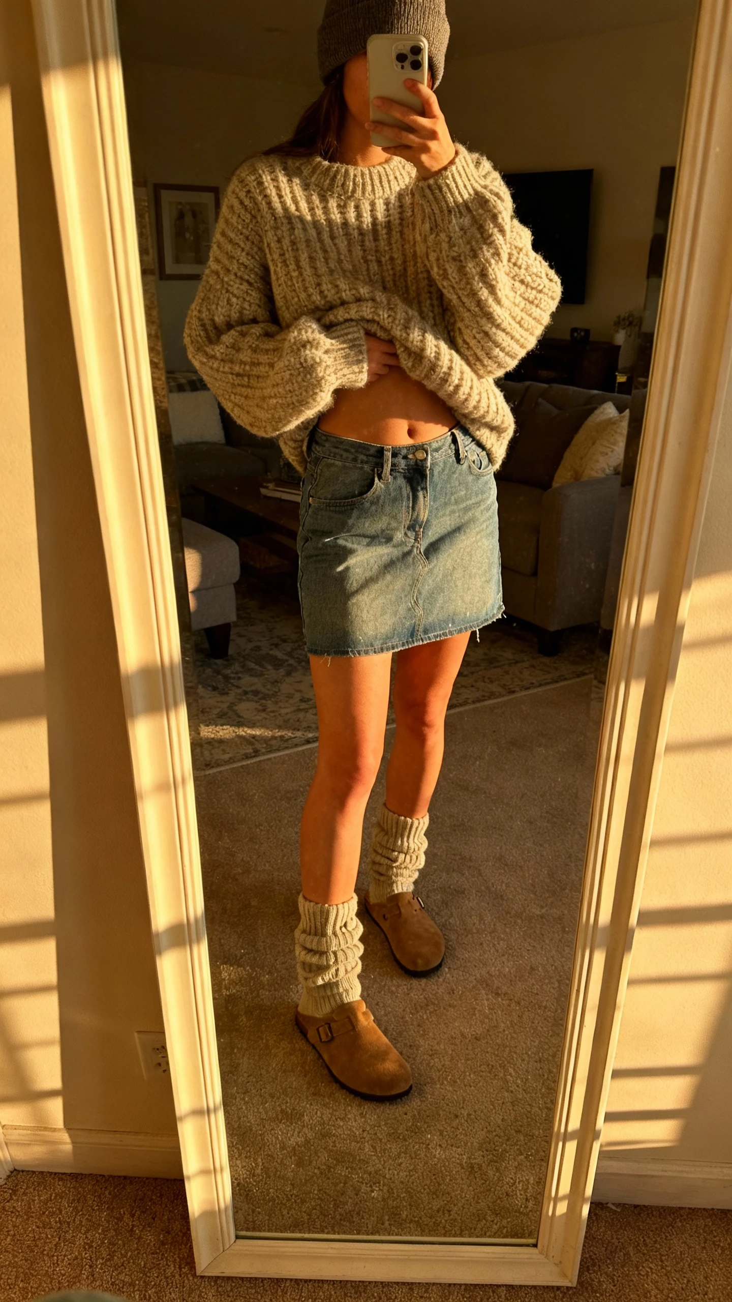 iPhone mirror selfie of a woman wearing an oversized chunky knit sweater half-tucked into a denim mini skirt, cozy slouch socks, suede clogs, and a beanie, face not visible, cropped at neck, living room mirror with warm afternoon light, iPhone photo quality.