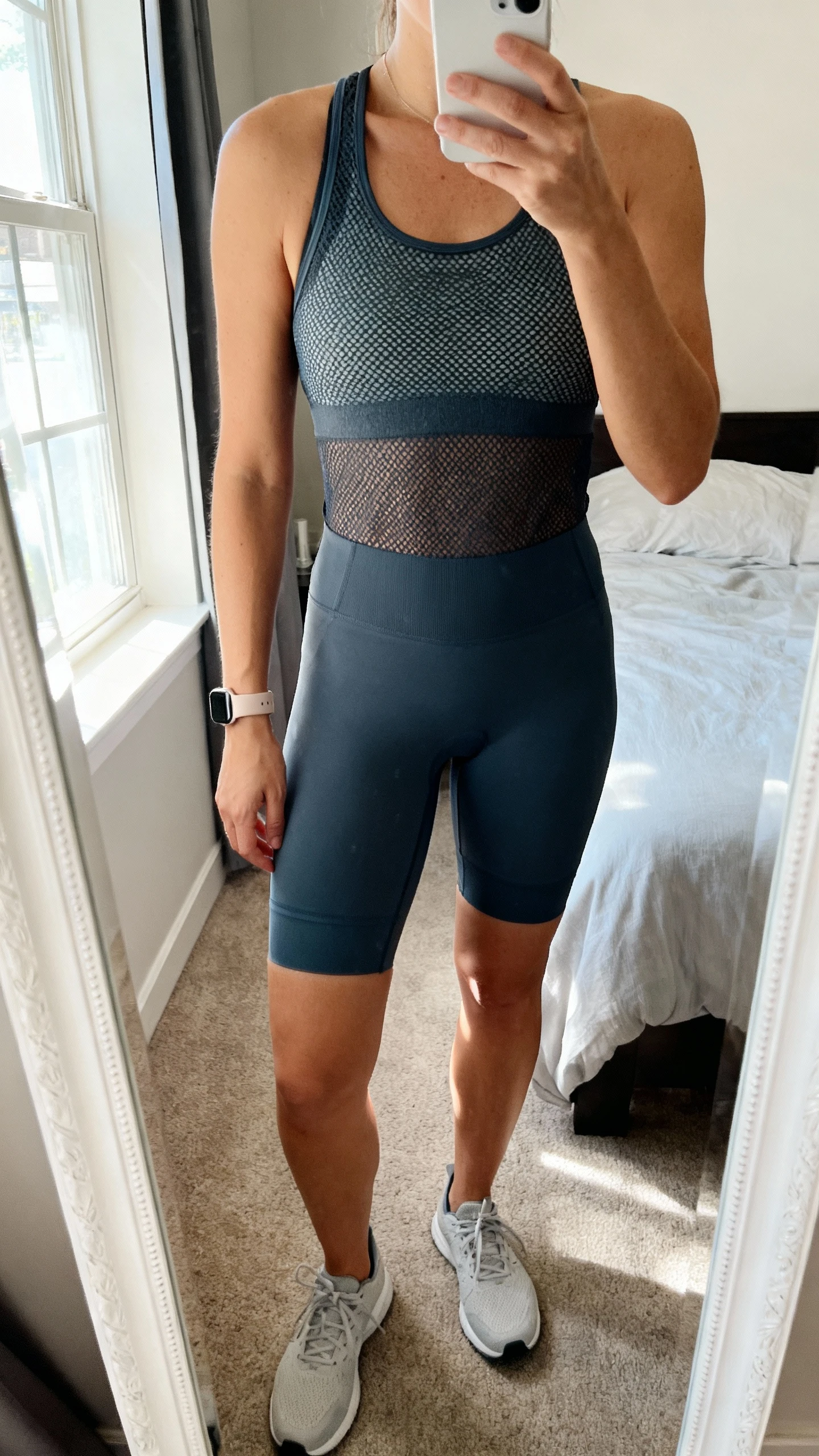 iPhone mirror selfie of a woman wearing an ultra-breathable mesh tank and matching bike shorts set, face not visible, cropped at neck, bedroom mirror with morning window light, minimal sneakers and a slim fitness watch, iPhone photo quality.