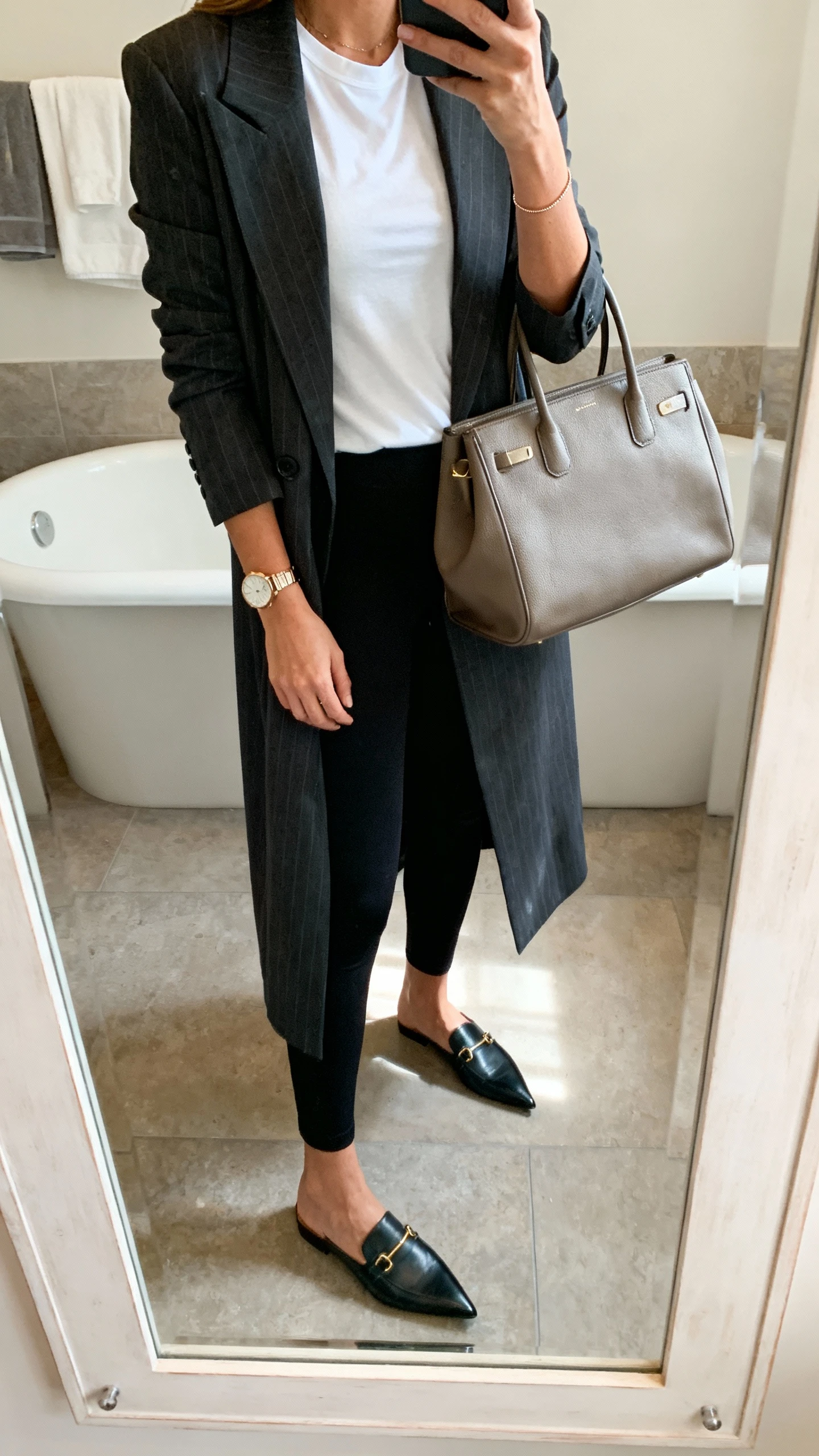 iPhone mirror selfie of a woman wearing ankle-length black leggings, longline tailored blazer, white fitted tee, pointed loafers, structured tote, delicate watch, face not visible, bathroom mirror, soft indoor lighting, iPhone photo quality