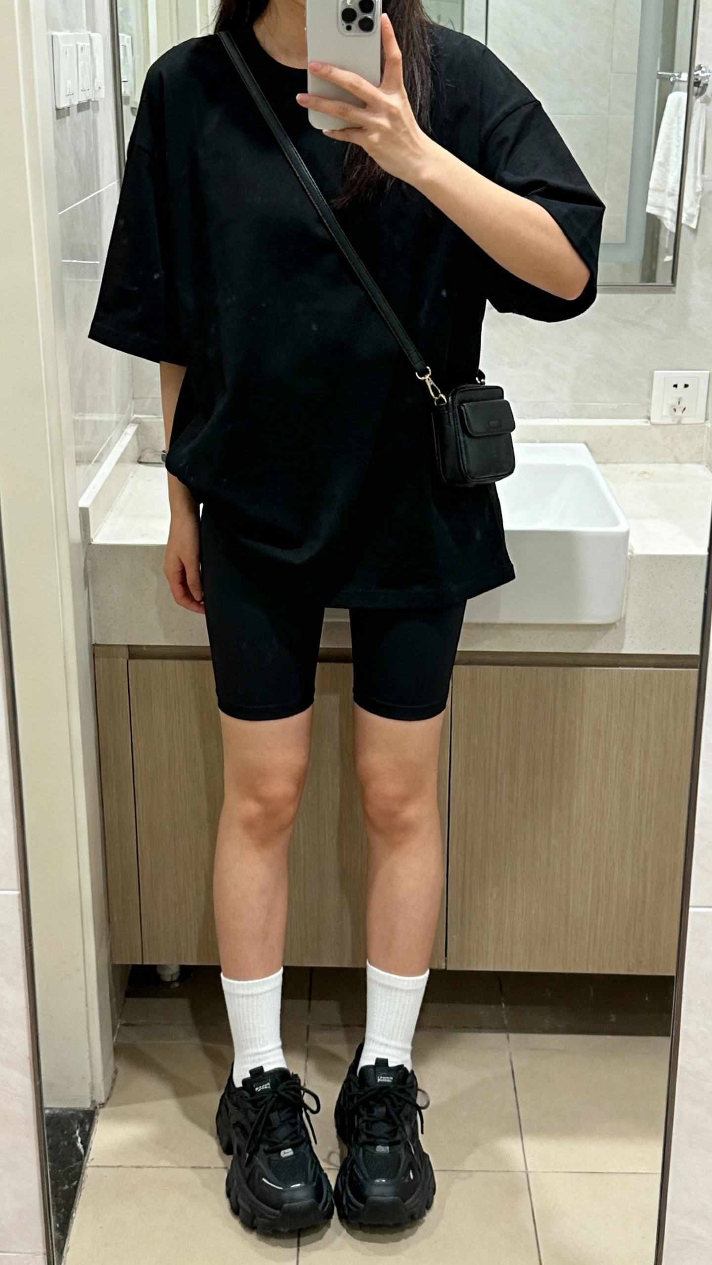 iPhone mirror selfie of a woman wearing black biker shorts and an oversized boxy black tee, face not visible, bathroom mirror with soft indoor lighting, white crew socks, chunky black trainers, and a small black crossbody, iPhone photo quality