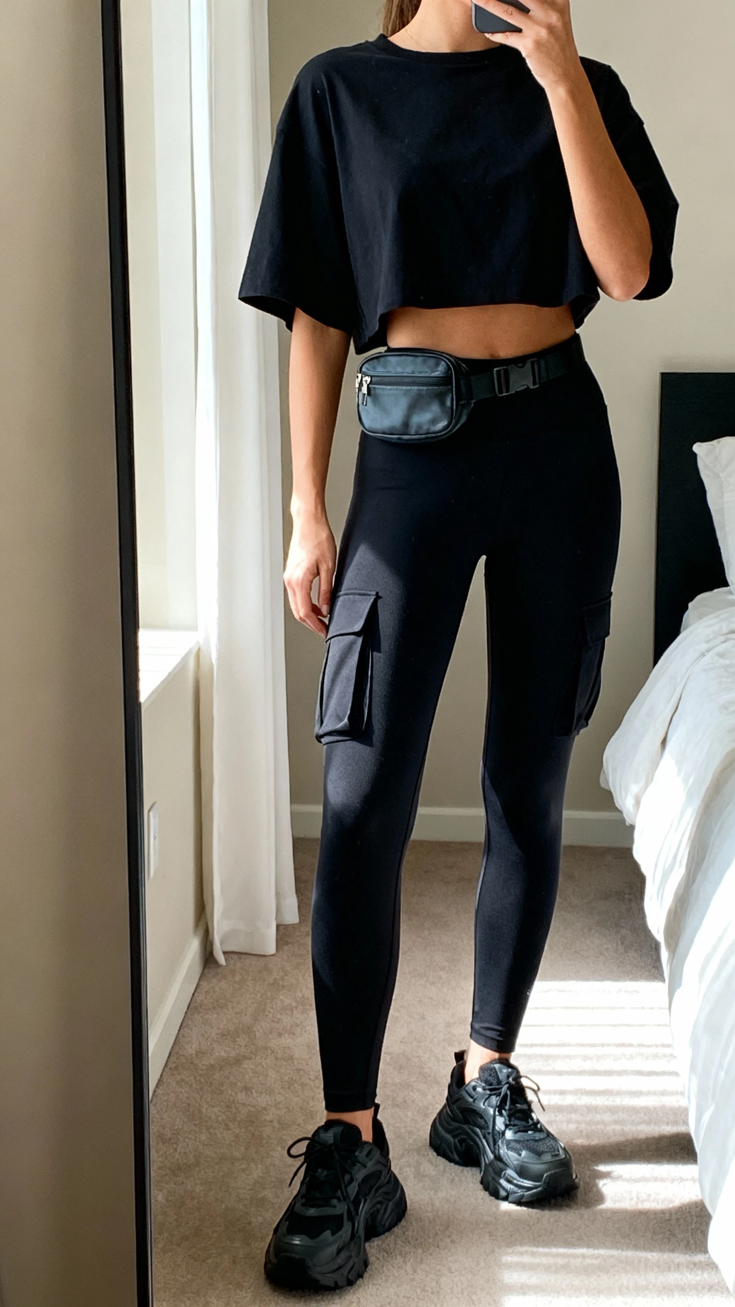 iPhone mirror selfie of a woman wearing black cargo-lite pocket leggings and a boxy black cropped tee with chunky sneakers, face not visible, cropped at neck, bedroom mirror with soft afternoon light, small belt bag at waist, iPhone photo quality.