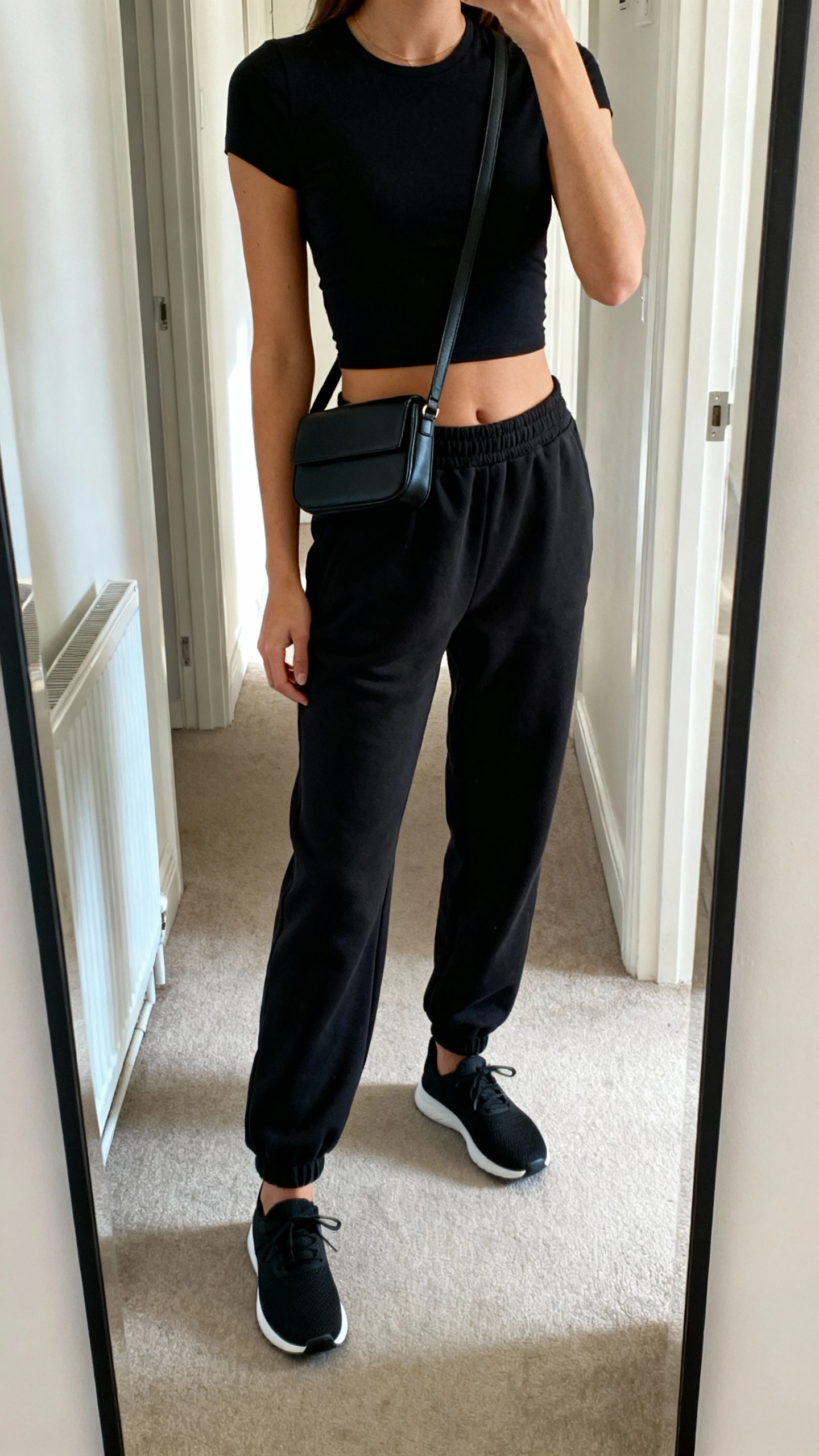 iPhone mirror selfie of a woman wearing black elevated joggers with a fitted black crop top, black low-profile sneakers, and a slim black crossbody belt bag, face not visible, cropped at shoulders, full-length hallway mirror, indoor daylight, iPhone photo quality.