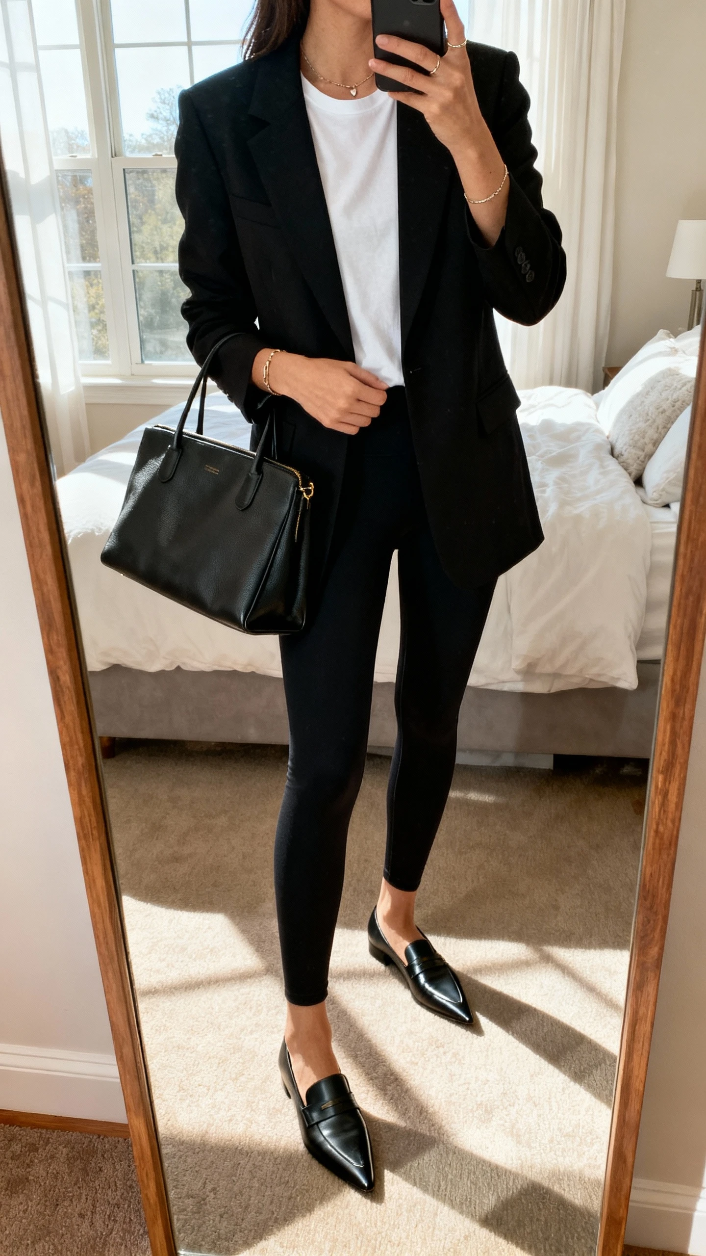 iPhone mirror selfie of a woman wearing black leggings, a tailored black blazer over a white tee, pointed loafers, structured tote bag, minimal jewelry, face not visible, cropped at neck, bedroom mirror with natural window light, iPhone photo quality