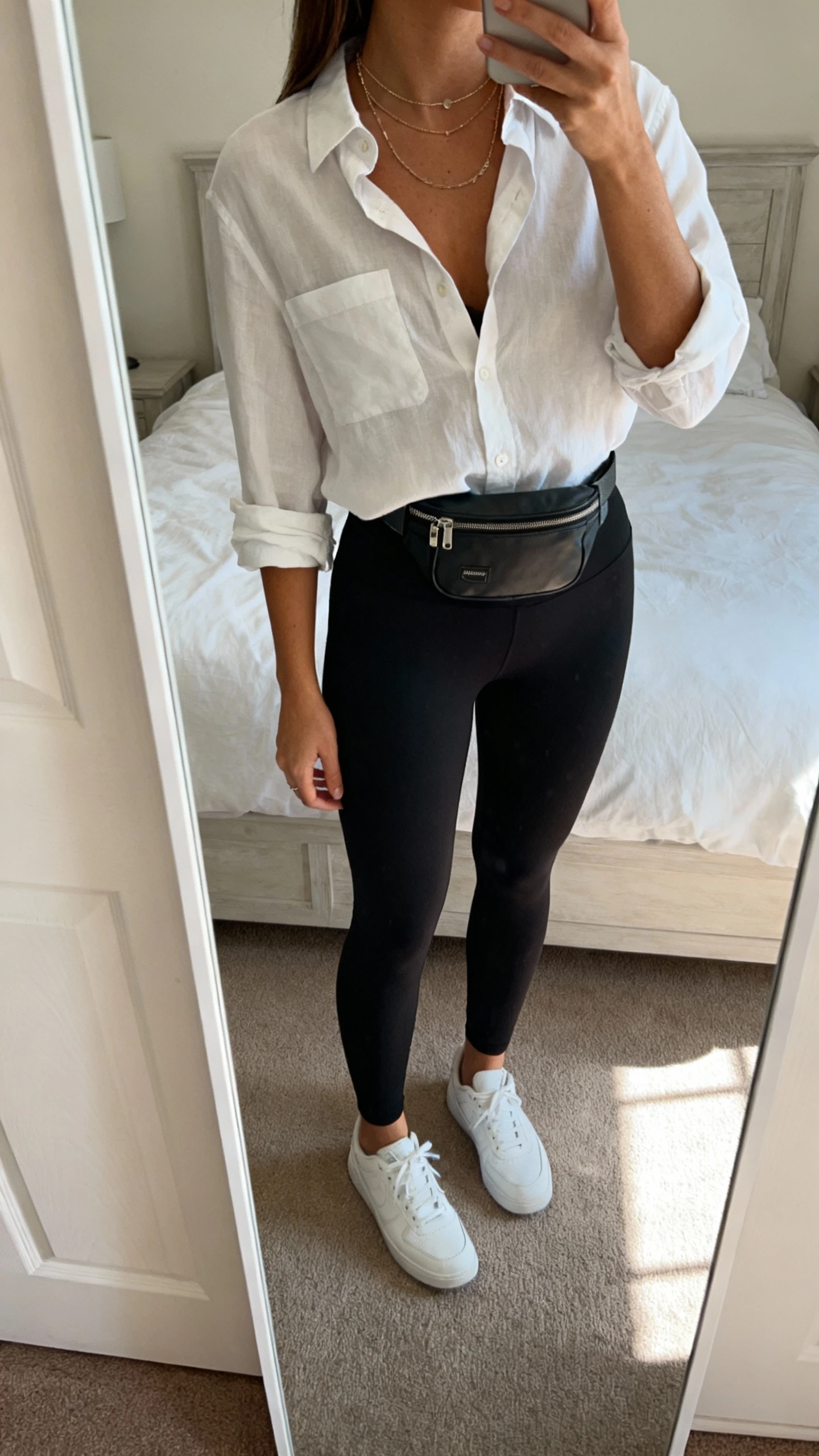 iPhone mirror selfie of a woman wearing black leggings, crisp button-up shirt slightly unbuttoned with sleeves rolled, belt bag at waist, white sneakers, layered delicate necklaces, face not visible, cropped at neck, bedroom mirror, daylight, iPhone photo quality