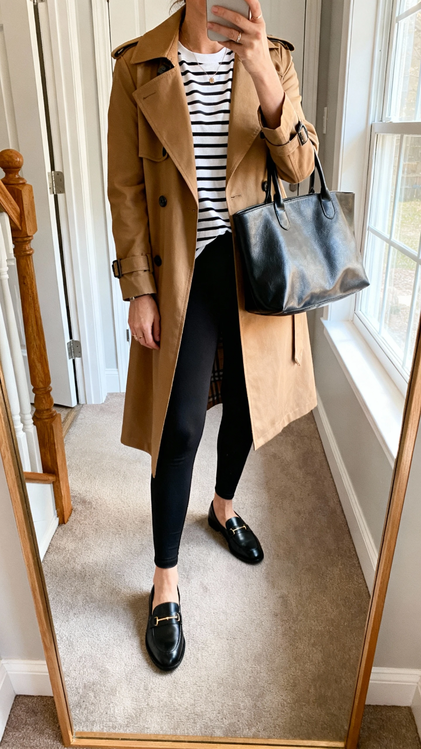 iPhone mirror selfie of a woman wearing black leggings, a classic camel trench coat, a black-and-white striped tee, black loafers, and a leather tote, face not visible, cropped at neck, entryway full-length mirror, morning window light, iPhone photo quality.