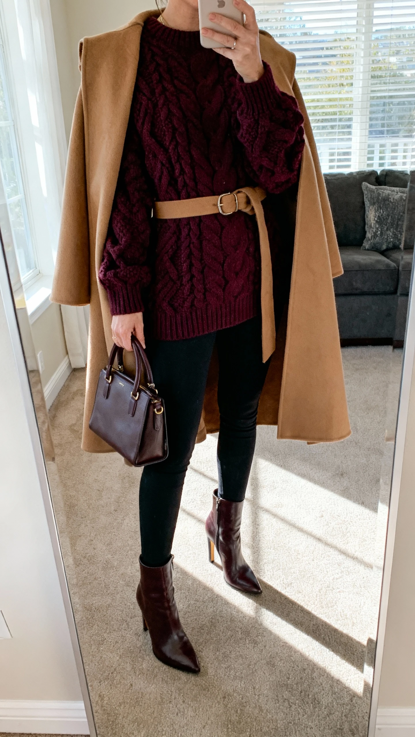 iPhone mirror selfie of a woman wearing black leggings, statement chunky knit sweater in deep burgundy, belted camel cape, heeled knee boots, top-handle bag, face not visible, cropped at shoulders, living-room full-length mirror, afternoon window light, iPhone photo quality.