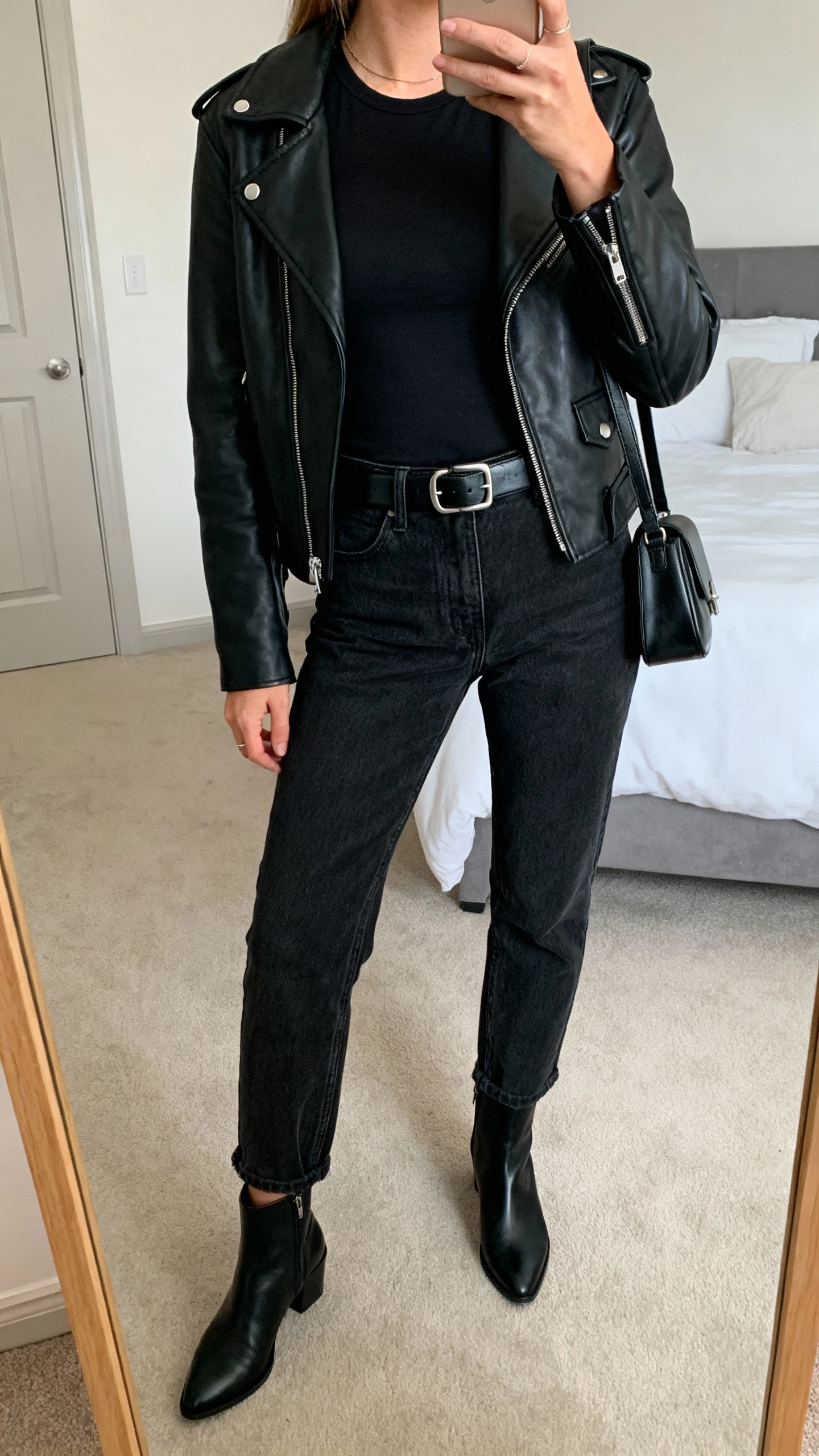 iPhone mirror selfie of a woman wearing black mom jeans, black fitted tee, black leather moto jacket, black leather belt with subtle buckle, black ankle boots, small black shoulder bag, face not visible, cropped at neck, bedroom mirror, soft indoor lighting, iPhone photo quality