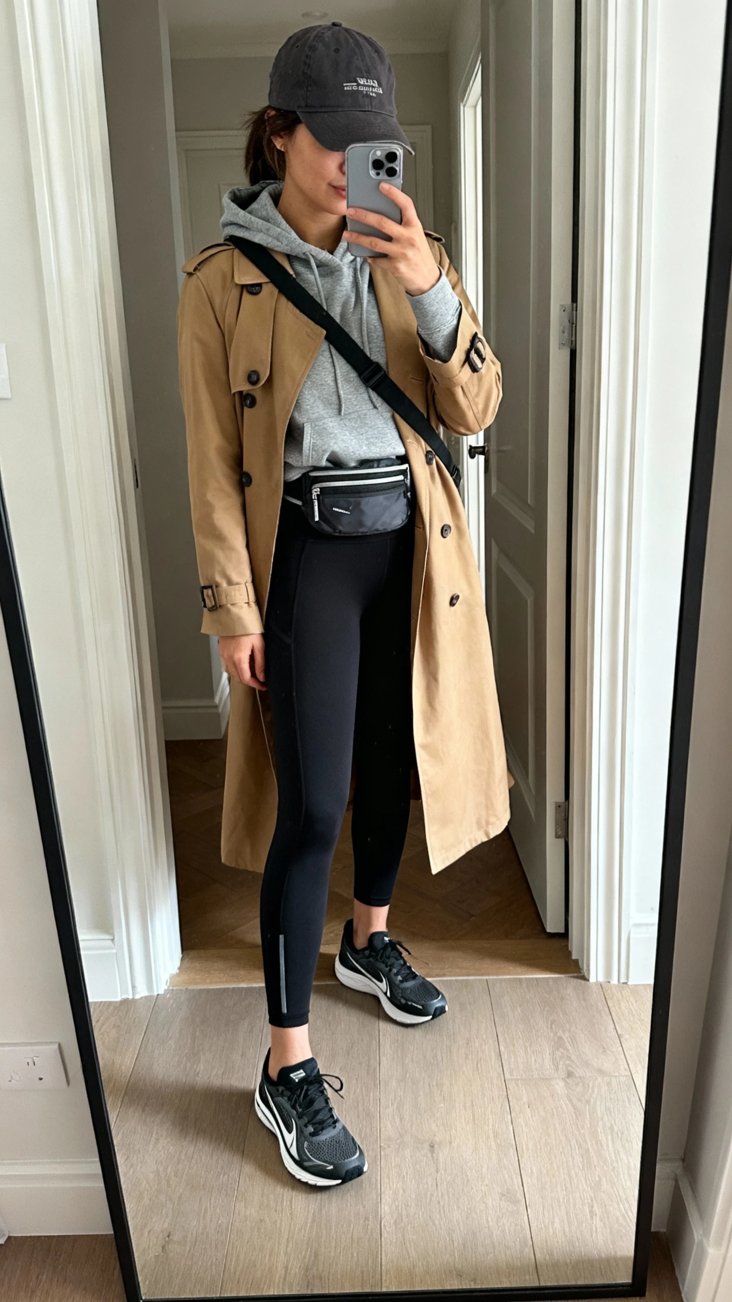 iPhone mirror selfie of a woman wearing black performance leggings, slate hoodie, classic tan trench, sleek running sneakers, baseball cap, crossbody belt bag, face not visible, cropped at neck, entryway full-length mirror, natural daylight, iPhone photo quality.