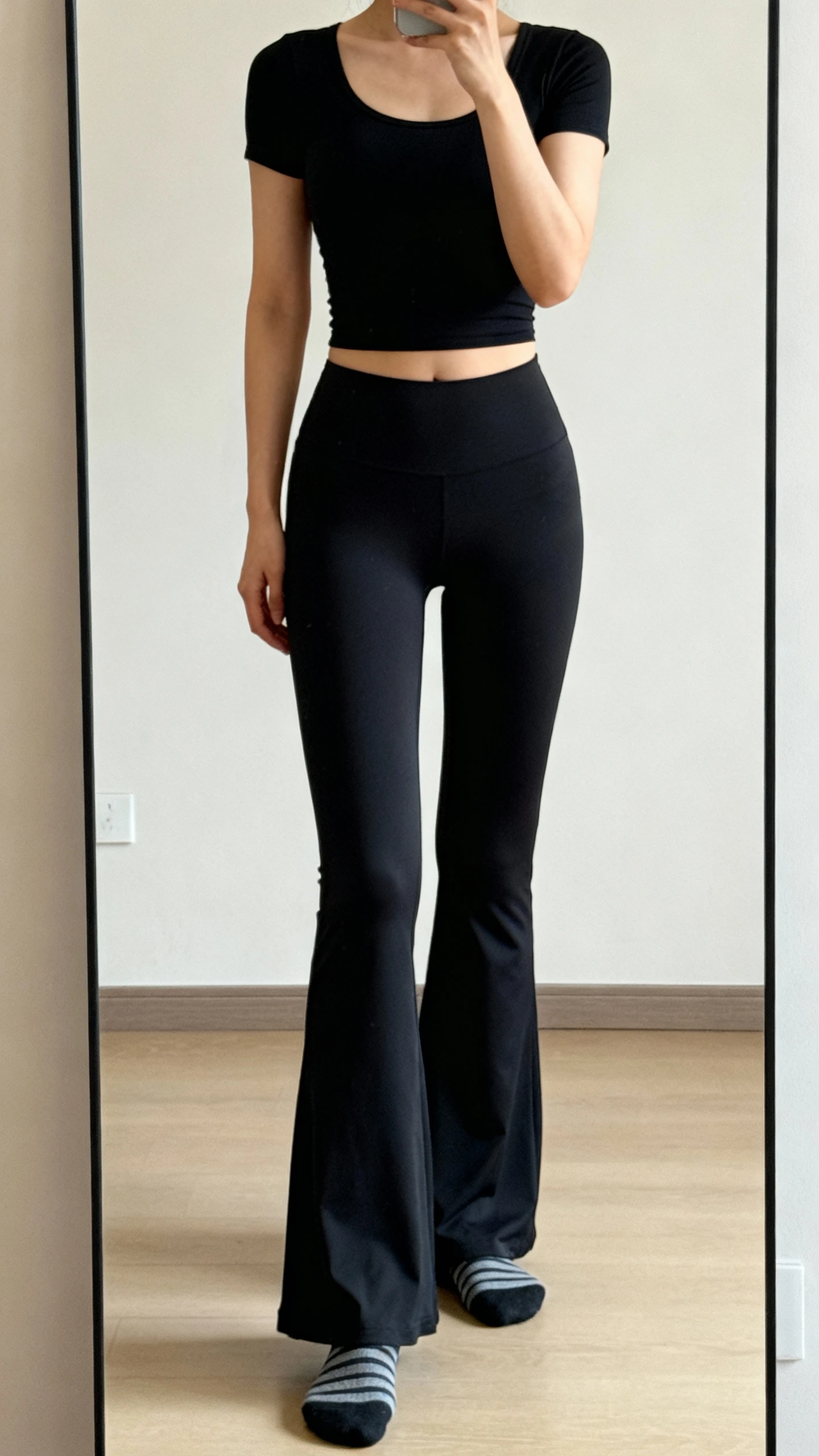 iPhone mirror selfie of a woman wearing black studio-ready flare leggings with a fitted black scoop-neck crop top and pilates socks, face not visible, cropped at neck, full-length mirror in a minimalist room, warm indoor light, iPhone photo quality.