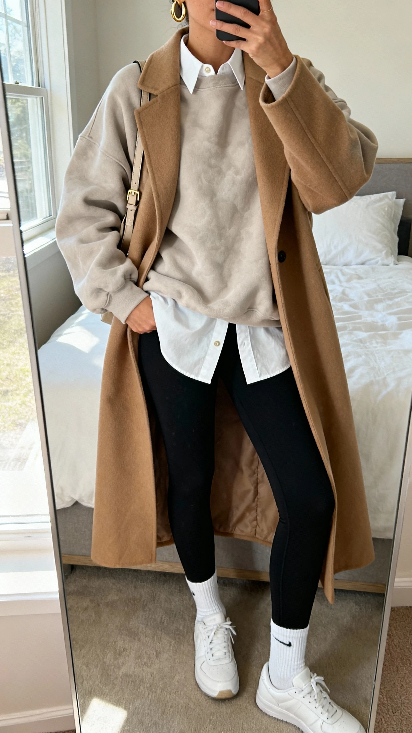 iPhone mirror selfie of a woman wearing cloud-soft oversized sweatshirt, black leggings, crisp white button-up peeking out, longline camel coat layered over, white crew socks, clean white sneakers, simple gold hoops, crossbody bag, face not visible, cropped at neck, bedroom mirror, natural window light, iPhone photo quality