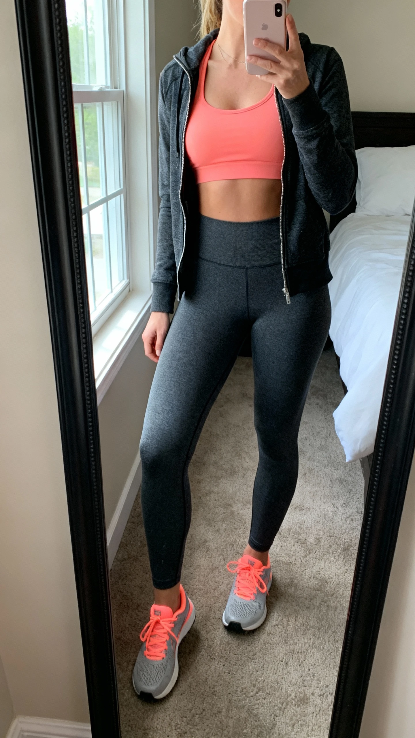 iPhone mirror selfie of a woman wearing coral sports bra, charcoal high-waist leggings, charcoal zip-up, and coral-accent trainers, face not visible, side angle, bedroom mirror with natural window light, iPhone photo quality.