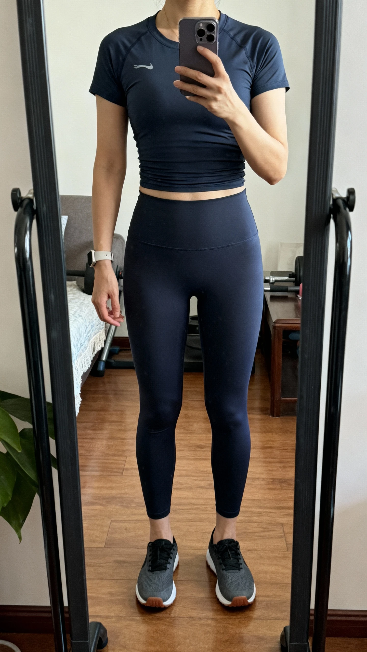 iPhone mirror selfie of a woman wearing flat, stable-sole lifting shoes, high-waisted leggings, and a fitted performance tee, face not visible, cropped at neck, gym-style full-length mirror at home, indoor lighting, iPhone photo quality