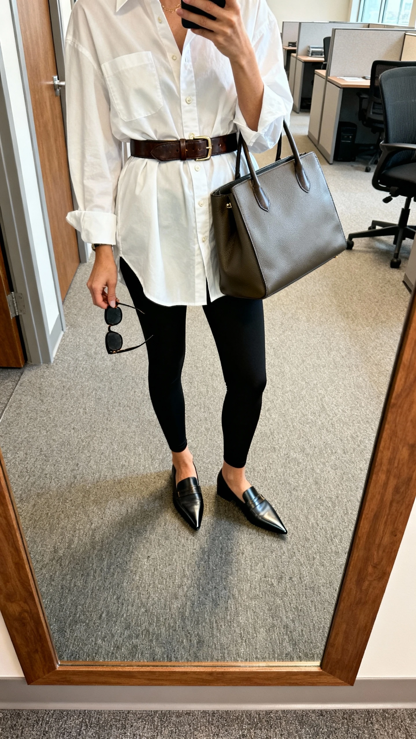 iPhone mirror selfie of a woman wearing matte black leggings, oversized crisp white button-down cinched with a leather belt, pointed loafers, structured tote, sunglasses in hand, face not visible, cropped at neck, office hallway mirror, natural daylight, iPhone photo quality.