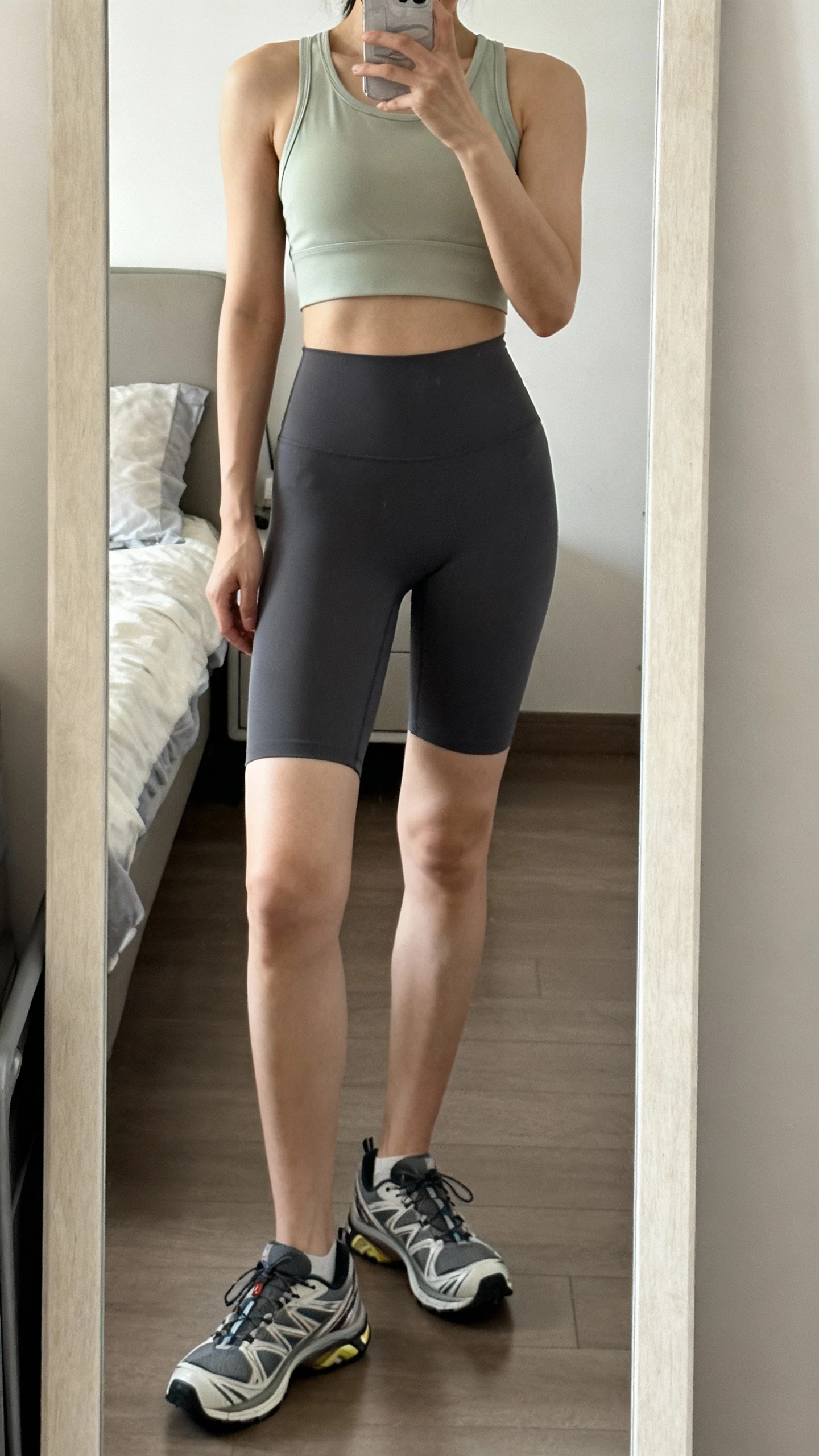 iPhone mirror selfie of a woman wearing mid-thigh compressive biker shorts that resist ride-up, a cropped performance tank, and trainers, face not visible, cropped at shoulders, bedroom mirror, soft indoor lighting, iPhone photo quality