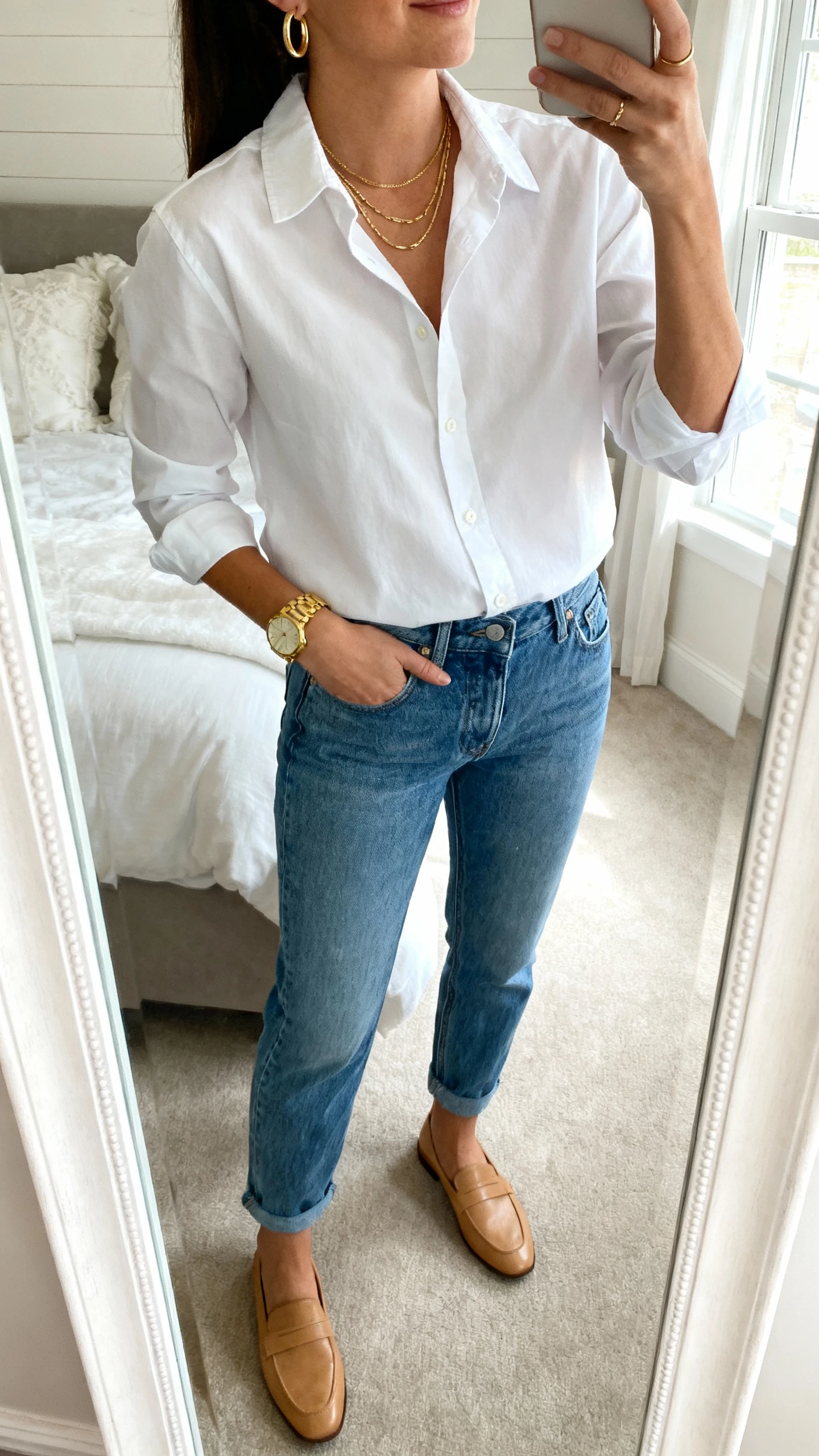 iPhone mirror selfie of a woman wearing mom jeans, a crisp white button-up shirt tucked in, slim gold hoop earrings, layered delicate gold necklaces, a gold watch, and tan loafers, face not visible, cropped at neck, bedroom mirror with natural window light, iPhone photo quality.