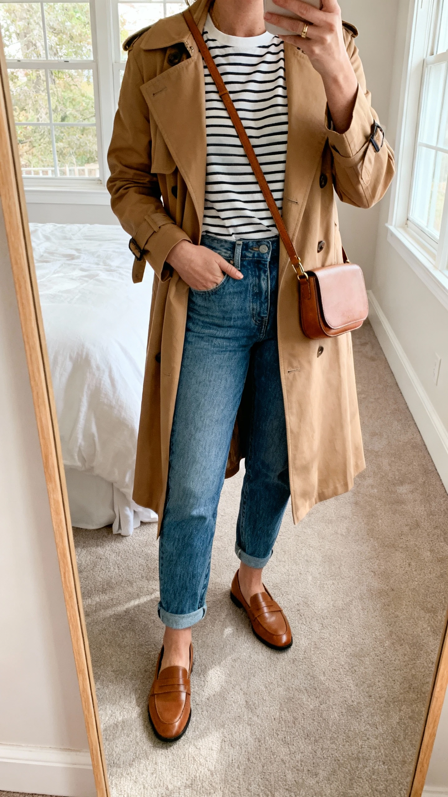 iPhone mirror selfie of a woman wearing mom jeans, a classic striped tee, and a camel trench coat with loafers and a simple leather crossbody, face not visible, cropped at neck, bedroom mirror, soft morning window light, iPhone photo quality