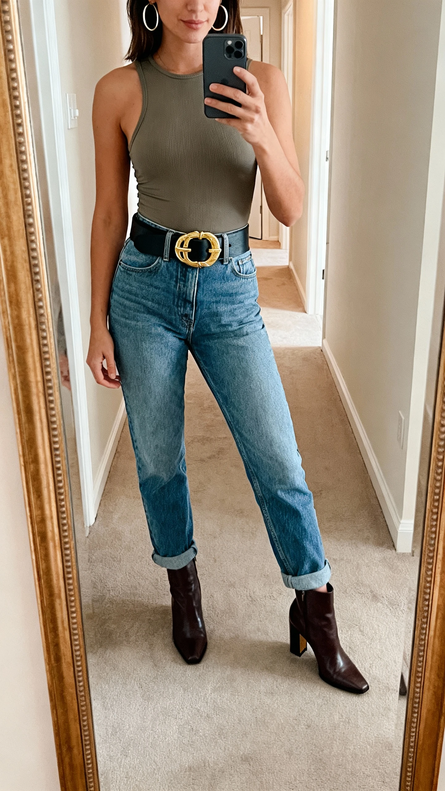 iPhone mirror selfie of a woman wearing mom jeans with a fitted bodysuit, a bold statement belt cinching the waist, hoop earrings, and heeled ankle boots, face not visible, cropped at neck, full-length mirror in hallway, indoor lighting, iPhone photo quality.