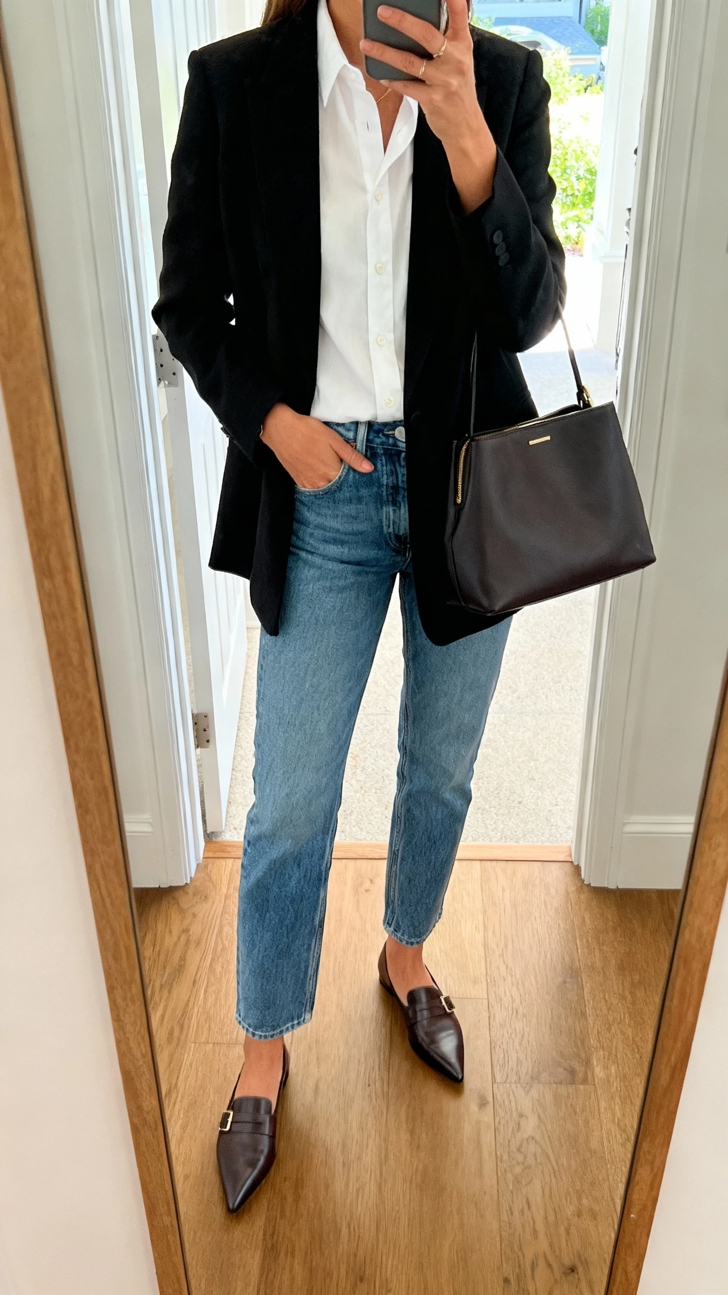 iPhone mirror selfie of a woman wearing mom jeans, a crisp white button-down, a sharp black blazer, pointed loafers, structured tote, face not visible, cropped at shoulders, entryway mirror, natural daylight, iPhone photo quality