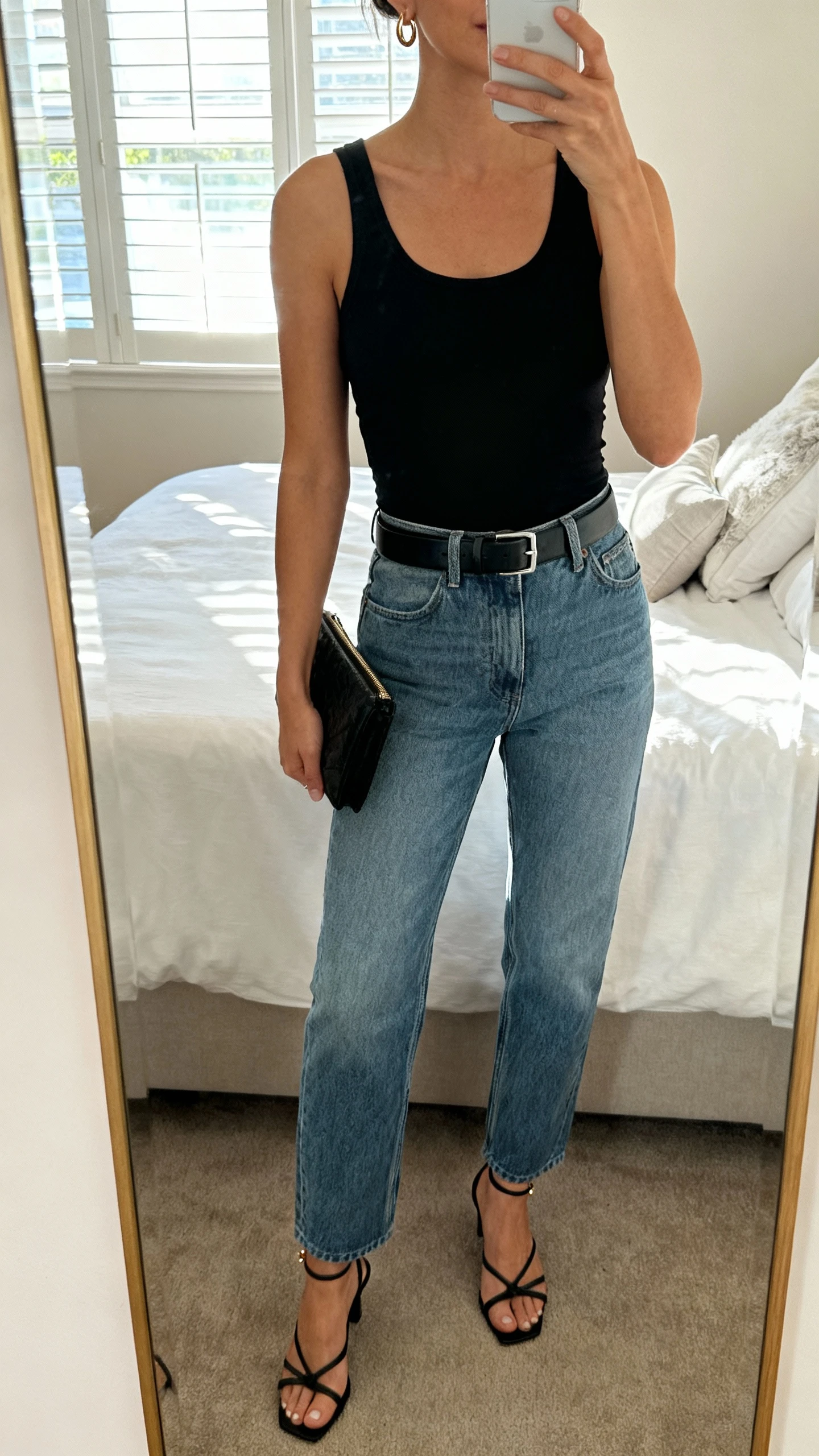 iPhone mirror selfie of a woman wearing mom jeans, a fitted black minimalist tank, sleek belt, and strappy heels with tiny hoop earrings and a clutch, face not visible, cropped at neck, bedroom mirror, natural window light, iPhone photo quality