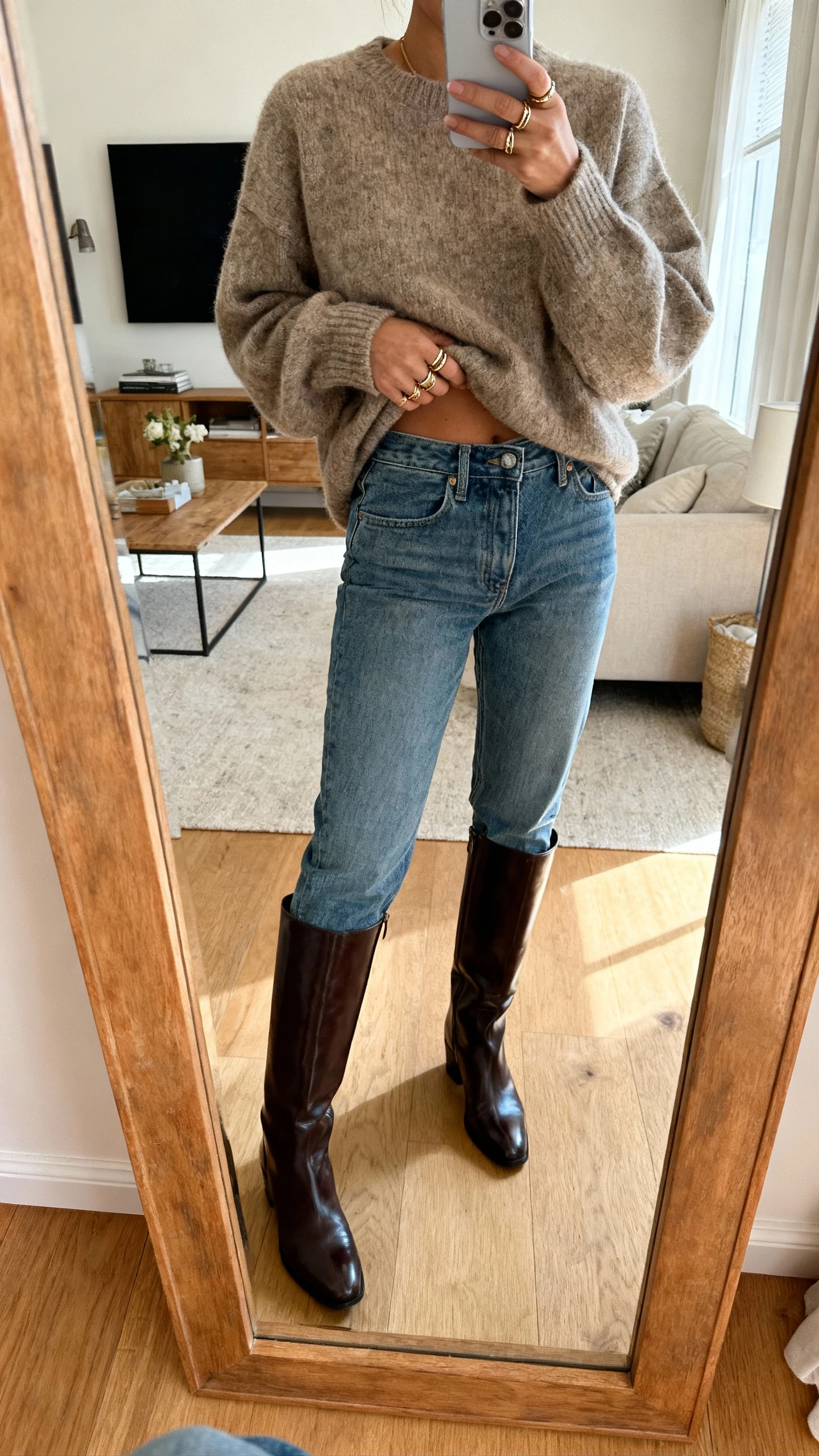 iPhone mirror selfie of a woman wearing mom jeans, a cozy oversized sweater half-tucked, sleek knee-high boots, and layered rings, face not visible, cropped at shoulders, living room mirror, warm indoor lighting, iPhone photo quality.