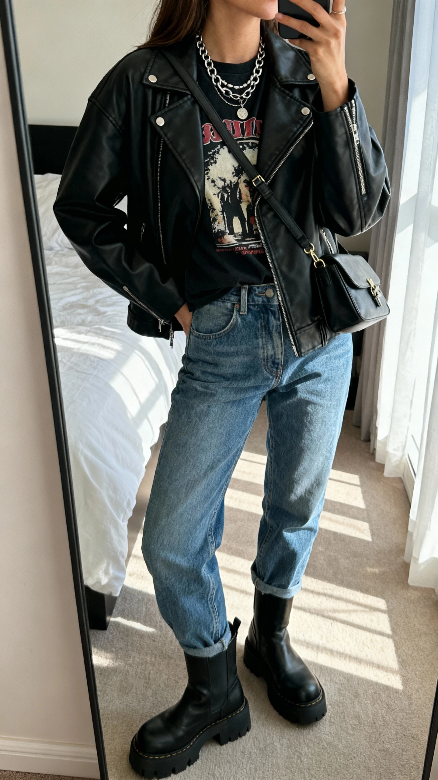 iPhone mirror selfie of a woman wearing mom jeans, a black leather jacket over a graphic tee, chunky boots, layered chain necklace and crossbody bag, face not visible, side angle, bedroom mirror, afternoon window light, iPhone photo quality