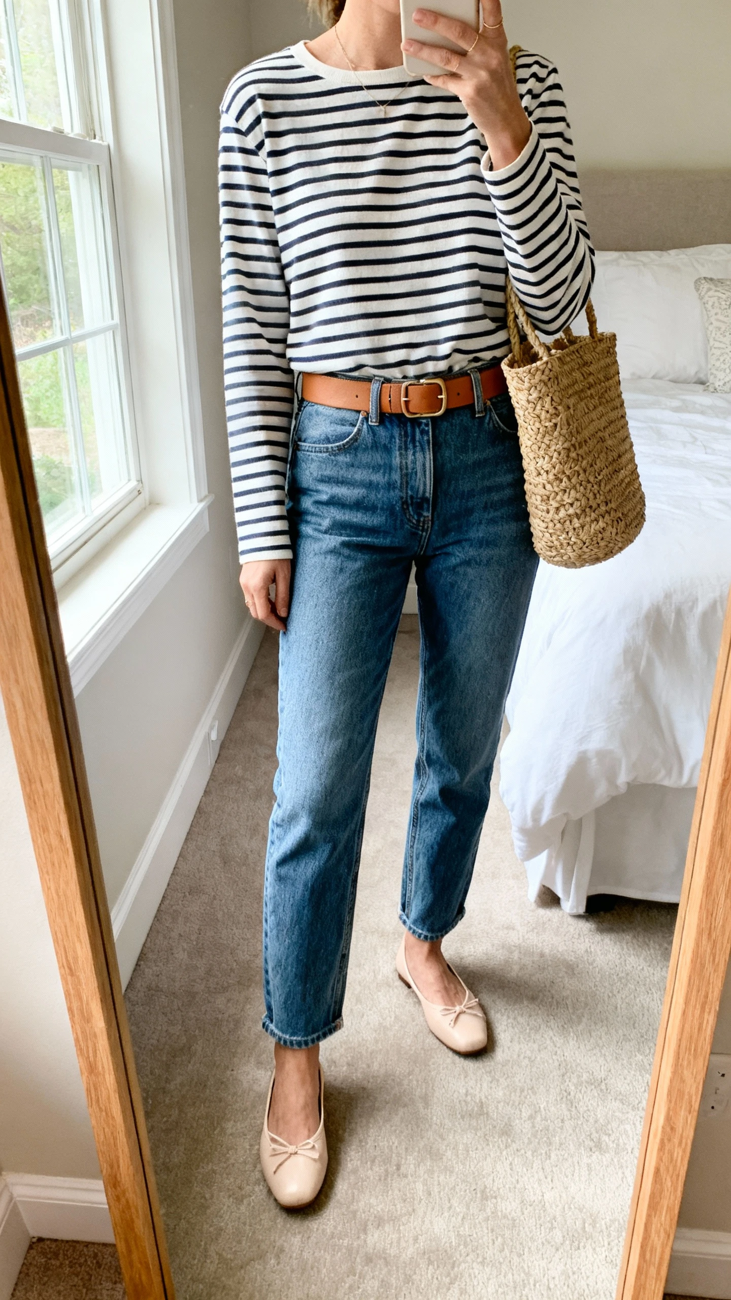 iPhone mirror selfie of a woman wearing mom jeans, a Breton striped long-sleeve top tucked in, ballet flats, a tan belt, and a woven tote, face not visible, cropped at neck, bedroom mirror, soft window light, iPhone photo quality.