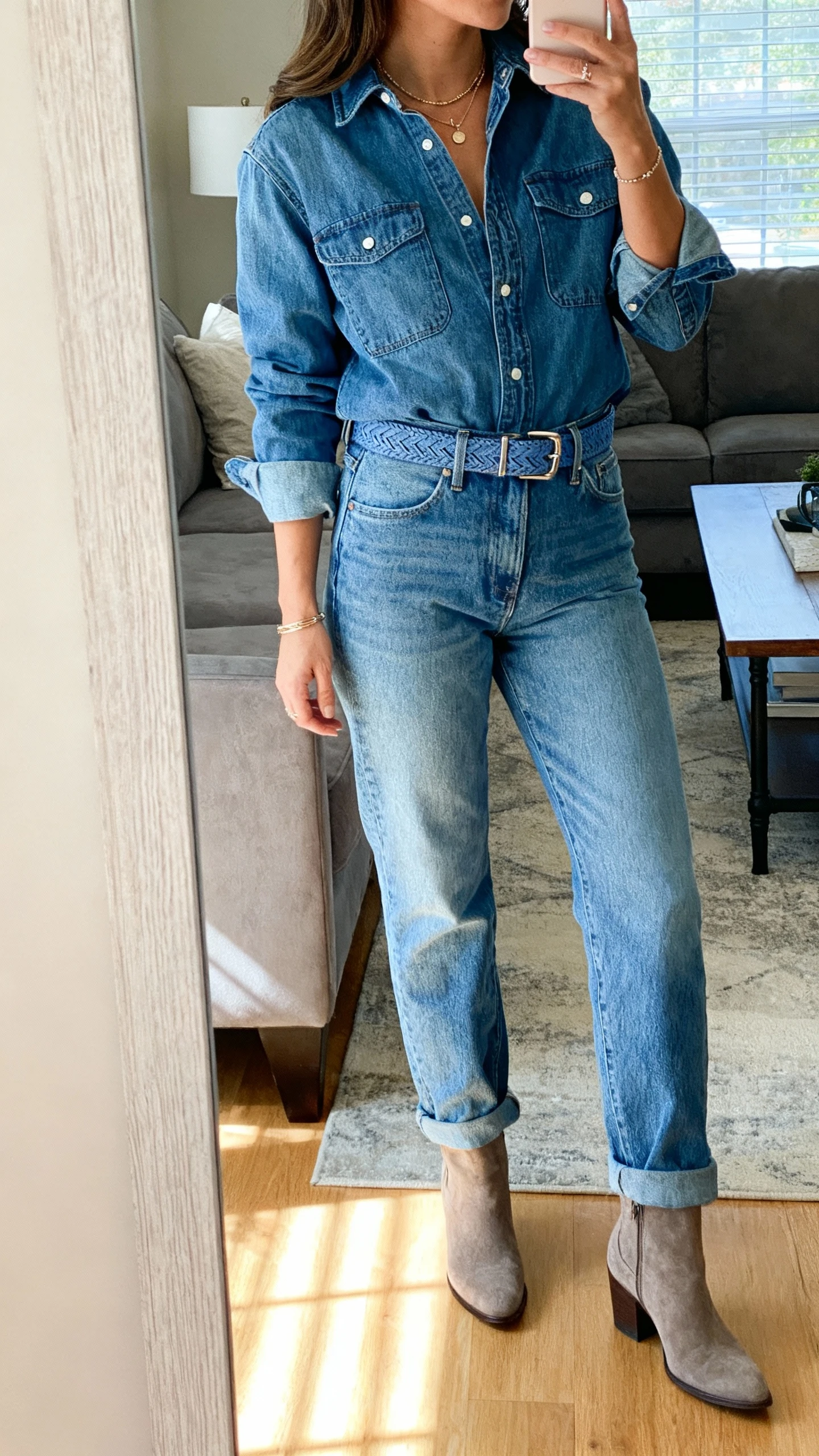 iPhone mirror selfie of a woman wearing monochrome denim-on-denim: mom jeans with a matching denim shirt/jacket, tonal belt, ankle boots, understated jewelry, face not visible, side angle, living room mirror, bright indoor light, iPhone photo quality