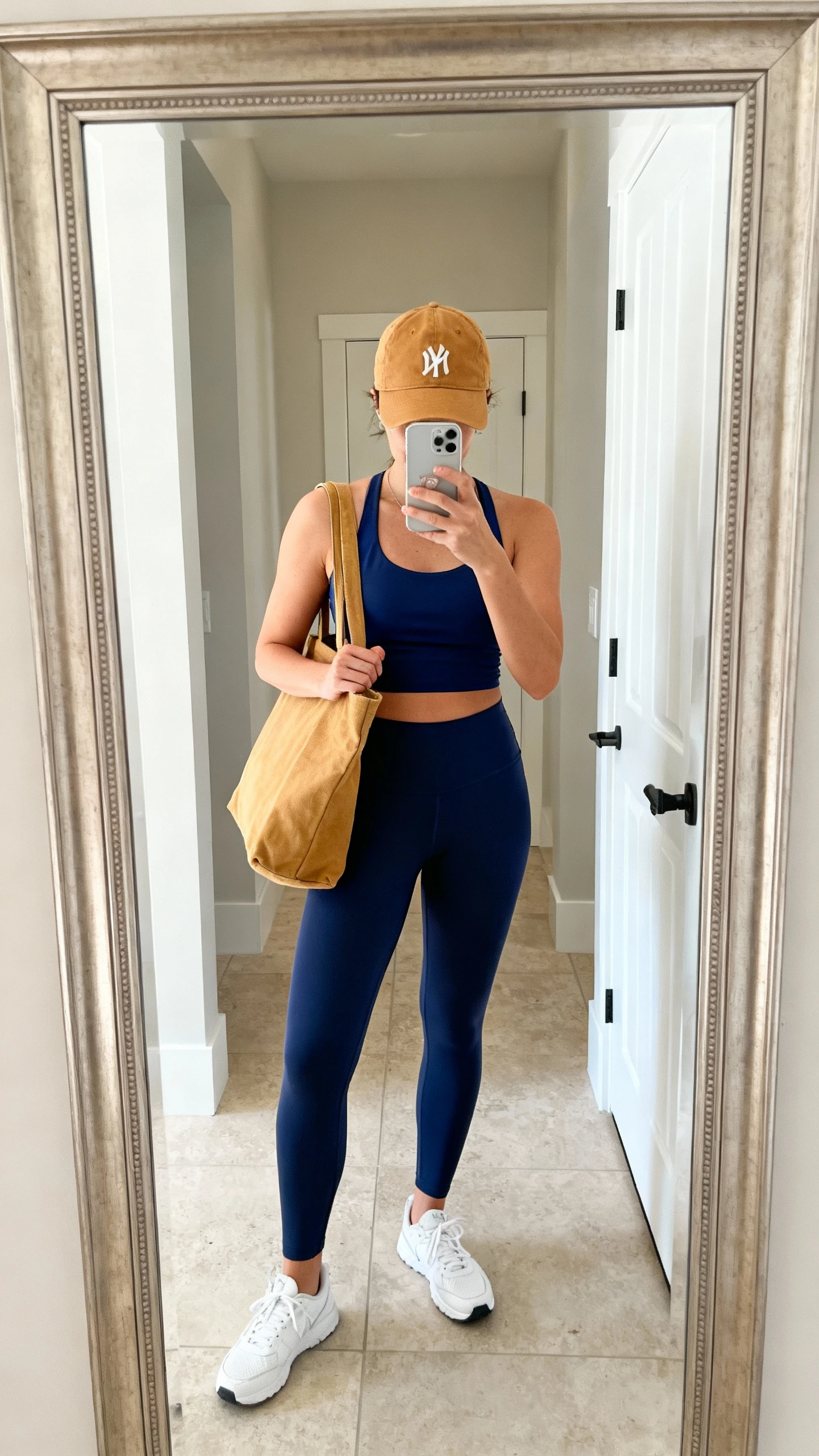 iPhone mirror selfie of a woman wearing navy leggings, a navy racerback top, white sneakers, and a tan baseball cap with a tan canvas tote, face not visible, cropped at neck, entryway mirror, bright natural light, iPhone photo quality.