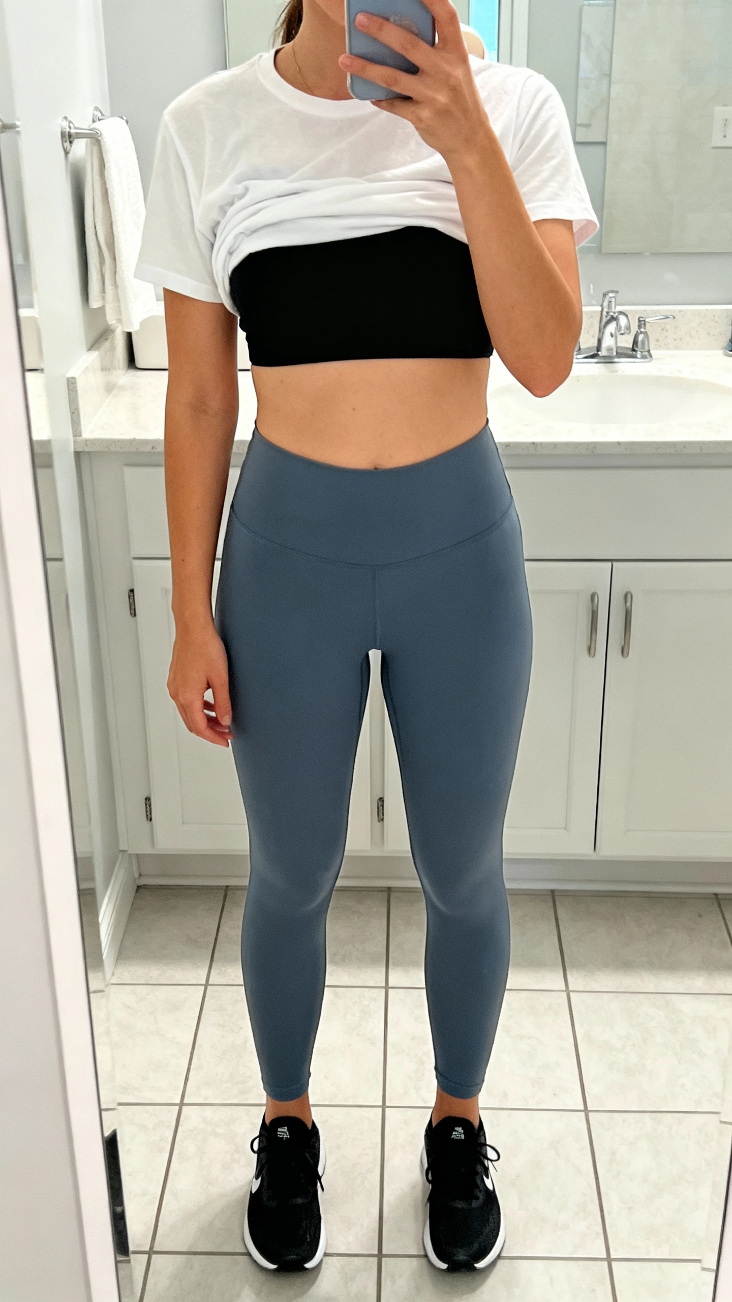 iPhone mirror selfie of a woman wearing slate blue leggings, a white cropped tee layered over a black sports bra, and black running shoes, face not visible, cropped at neck, bathroom mirror, bright indoor light, iPhone photo quality.