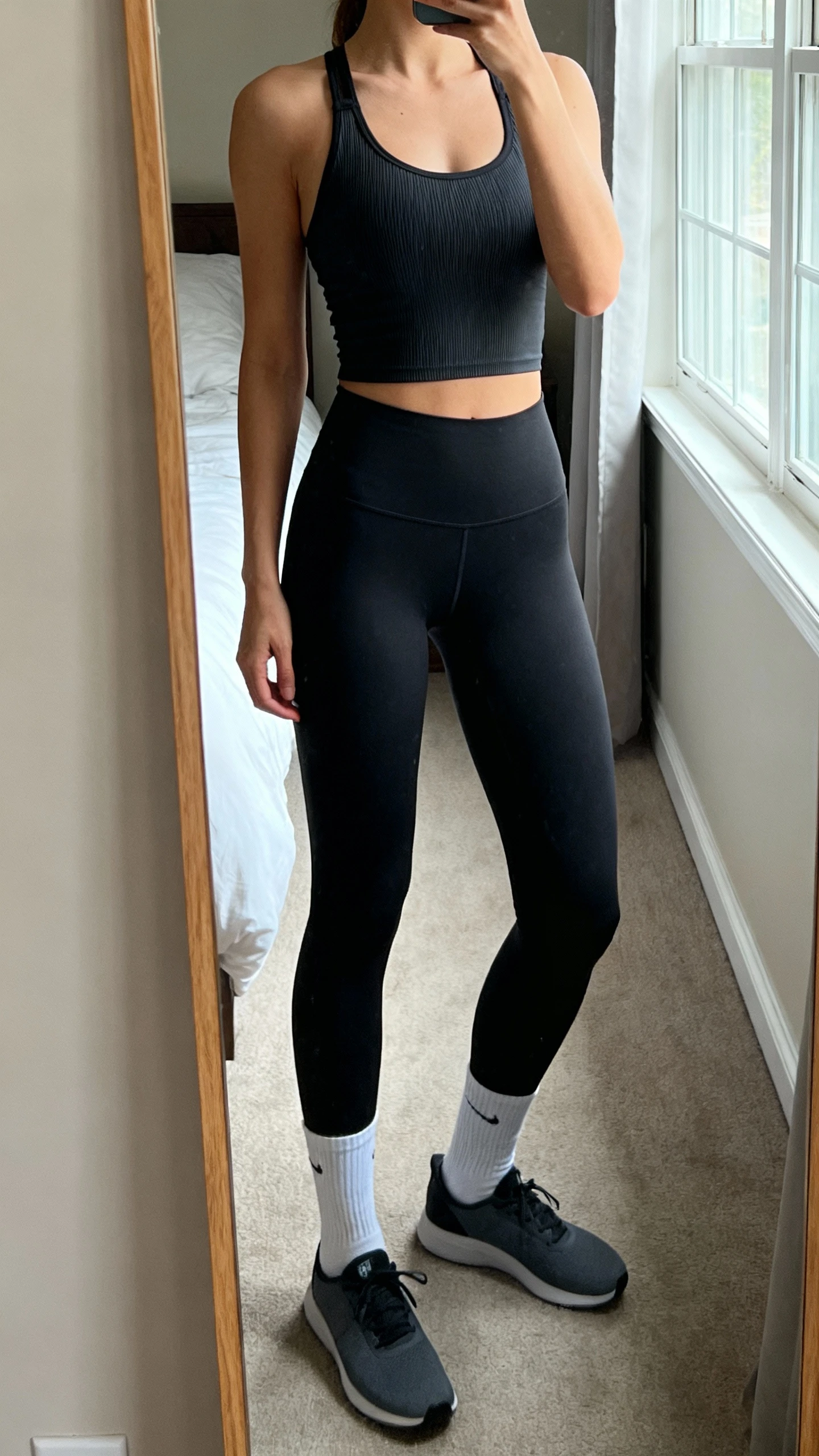 iPhone mirror selfie of a woman wearing squat-proof black high-waisted leggings, a compressive moisture-wicking racerback tank, ankle socks, and flat training sneakers, face not visible, cropped at neck, bedroom mirror with morning window light, iPhone photo quality