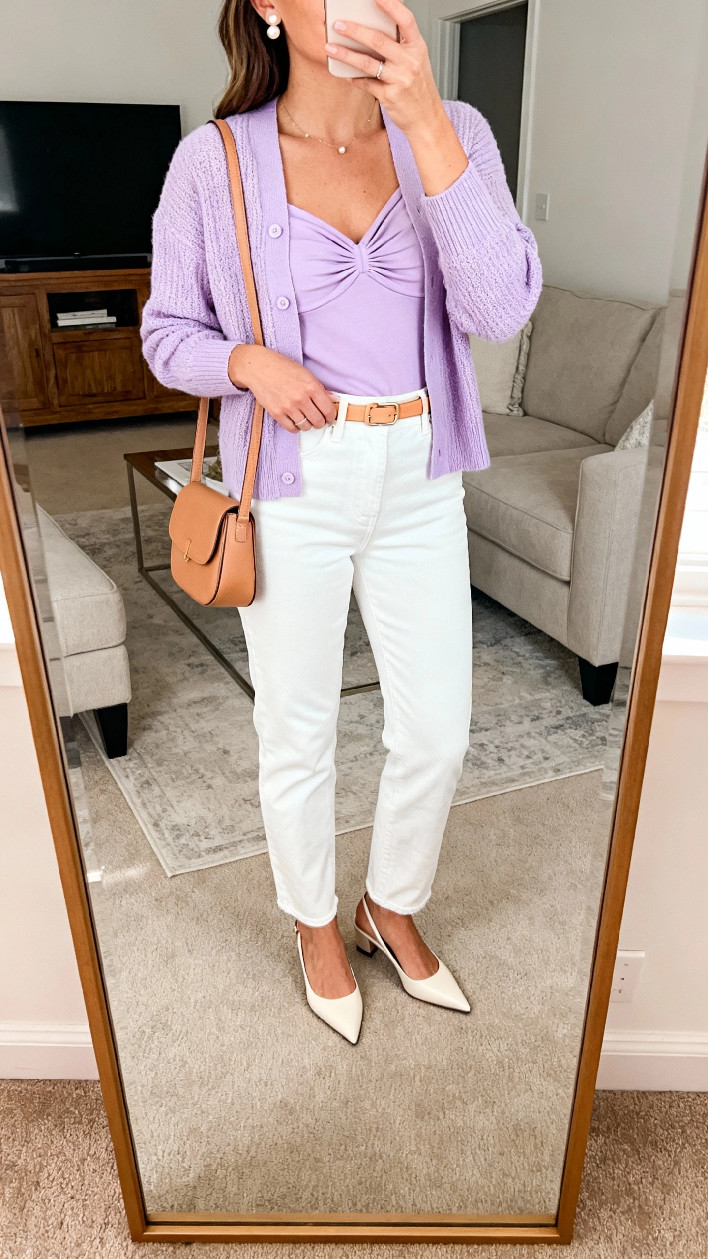 iPhone mirror selfie of a woman wearing straight-leg white jeans with a lilac cardigan and matching shell, pointed slingbacks, small tan shoulder bag, delicate pearl studs, thin camel belt, face not visible, cropped at shoulders, living room full-length mirror, natural daylight, iPhone photo quality.