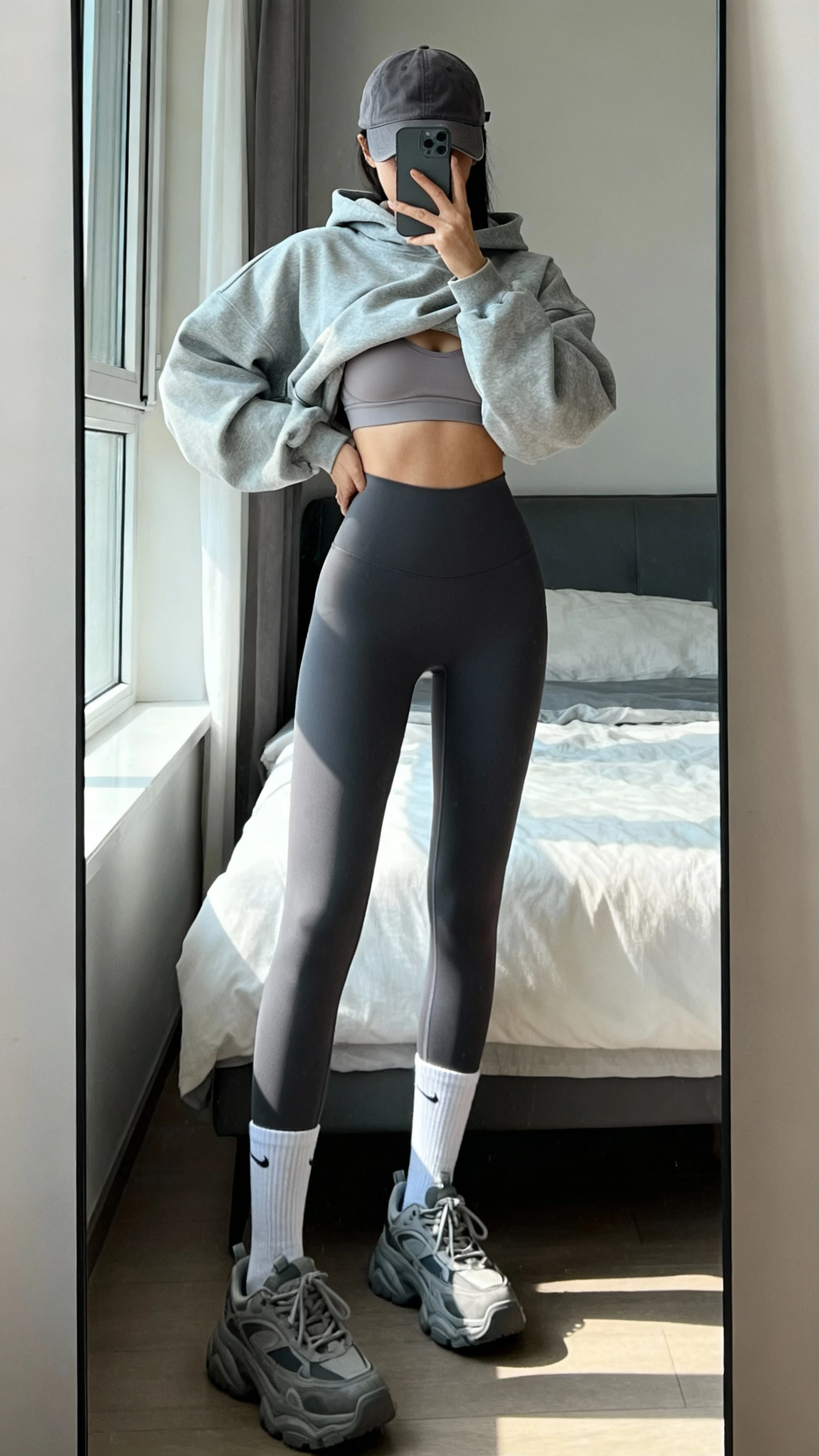 iPhone mirror selfie of a woman wearing streetwear-strong layering: oversized cropped hoodie over a supportive bra, compressive leggings, chunky trainers, calf socks, and a curved-brim cap, face not visible, cropped at shoulders, bedroom full-length mirror, natural window light, iPhone photo quality