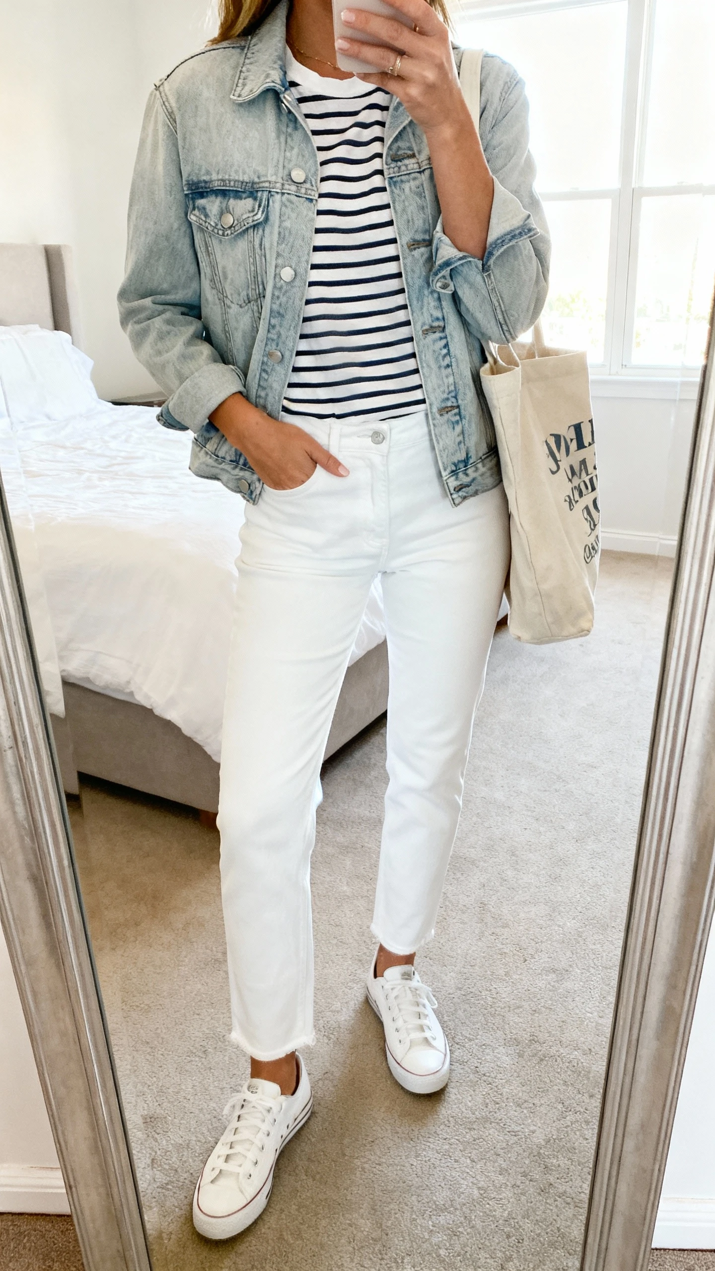 iPhone mirror selfie of a woman wearing white straight-leg jeans, a light-wash denim jacket over a striped tee, white sneakers, and a canvas tote, face not visible, cropped at shoulders, bedroom mirror, bright window light, iPhone photo quality.