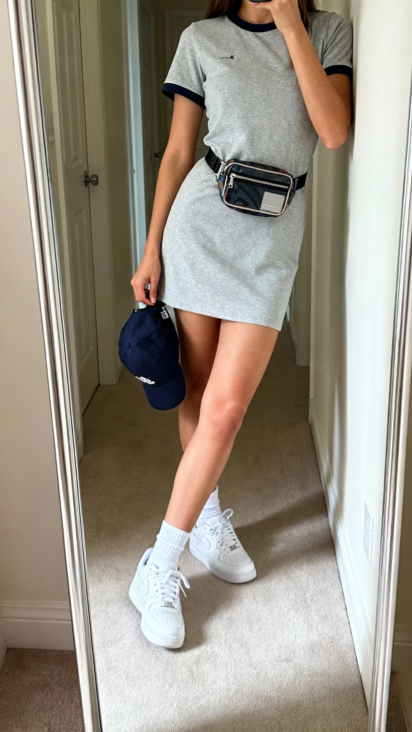 iPhone side-angle hallway mirror selfie of a woman wearing a sporty-chic tee dress with clean white sneakers, ankle socks, baseball cap in hand, and a small crossbody belt bag, face not visible, cropped at shoulders, natural daylight, iPhone photo quality.