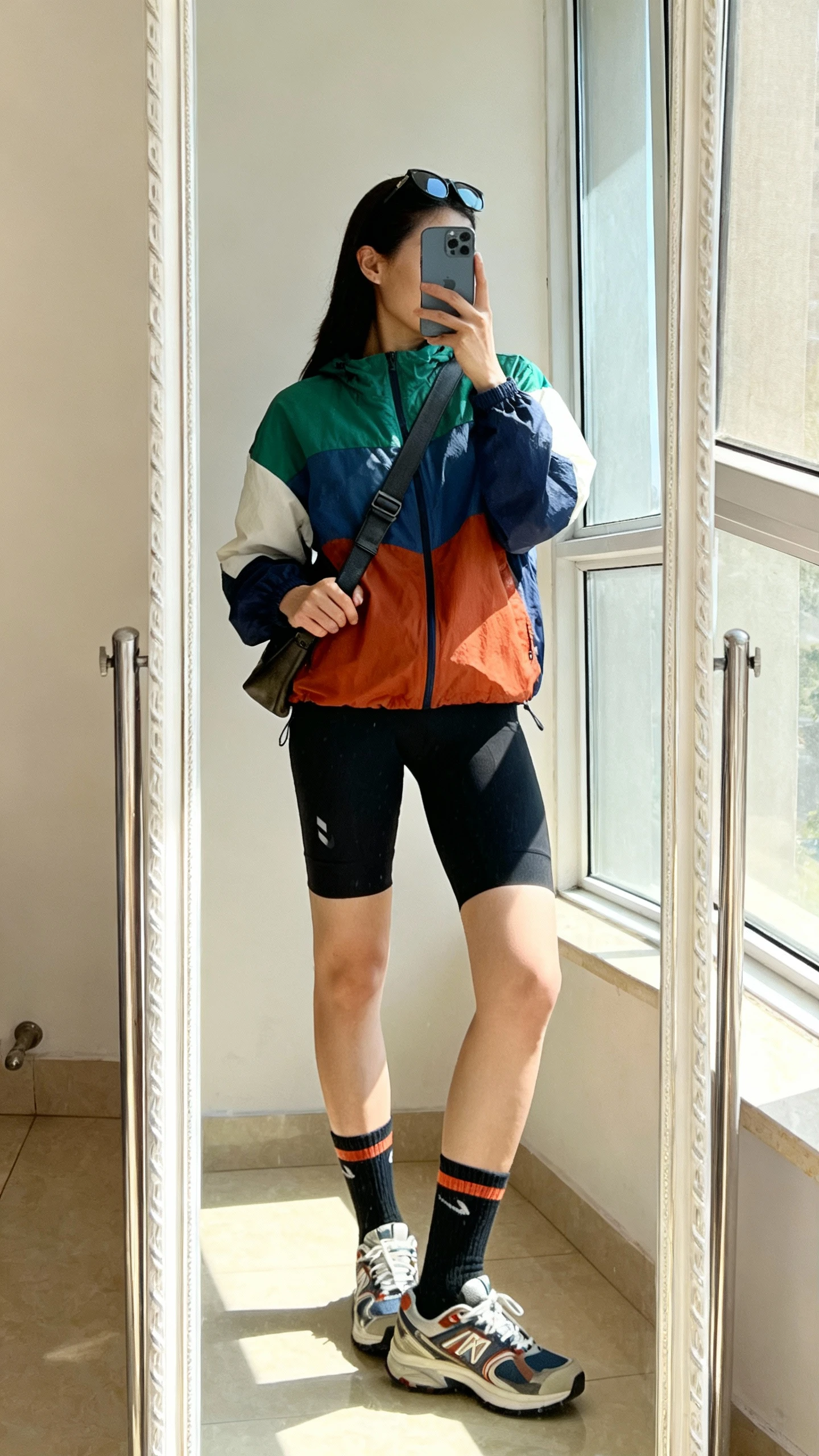 iPhone side-angle mirror selfie of a woman in a color-block windbreaker paired with black bike shorts, sporty crew socks, retro trainers, crossbody sling bag, sunglasses on head, face not visible, full-length mirror, bright daylight near window, iPhone photo quality.