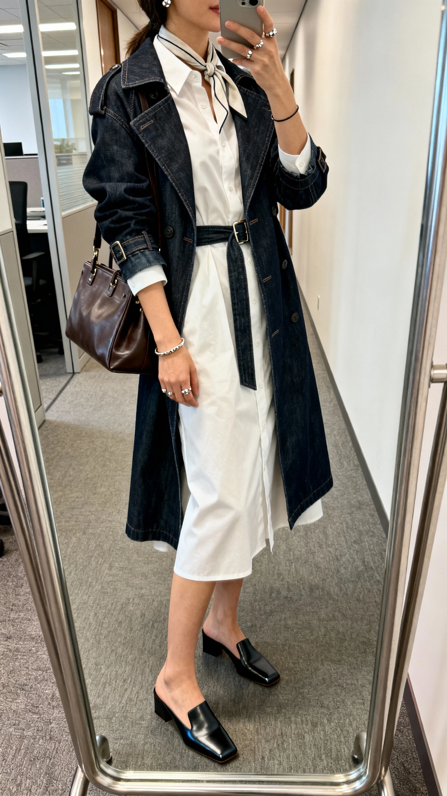 iPhone side-angle mirror selfie of a woman in a crisp midi belted shirt dress with a dark-wash denim trench, sculptural square-toe flats, a structured leather tote, statement studs and a slim bangle (optional thin silk scarf at the neck), face not visible, office hallway mirror, indoor lighting, iPhone photo quality.