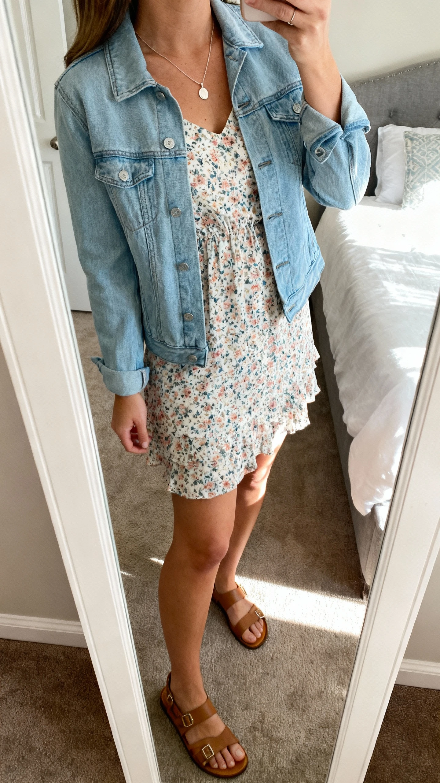 iPhone side-angle mirror selfie of a woman in a floral day dress with a light blue denim jacket, delicate pendant necklace, and sandals, face not visible, bedroom mirror, natural daylight, iPhone photo quality.