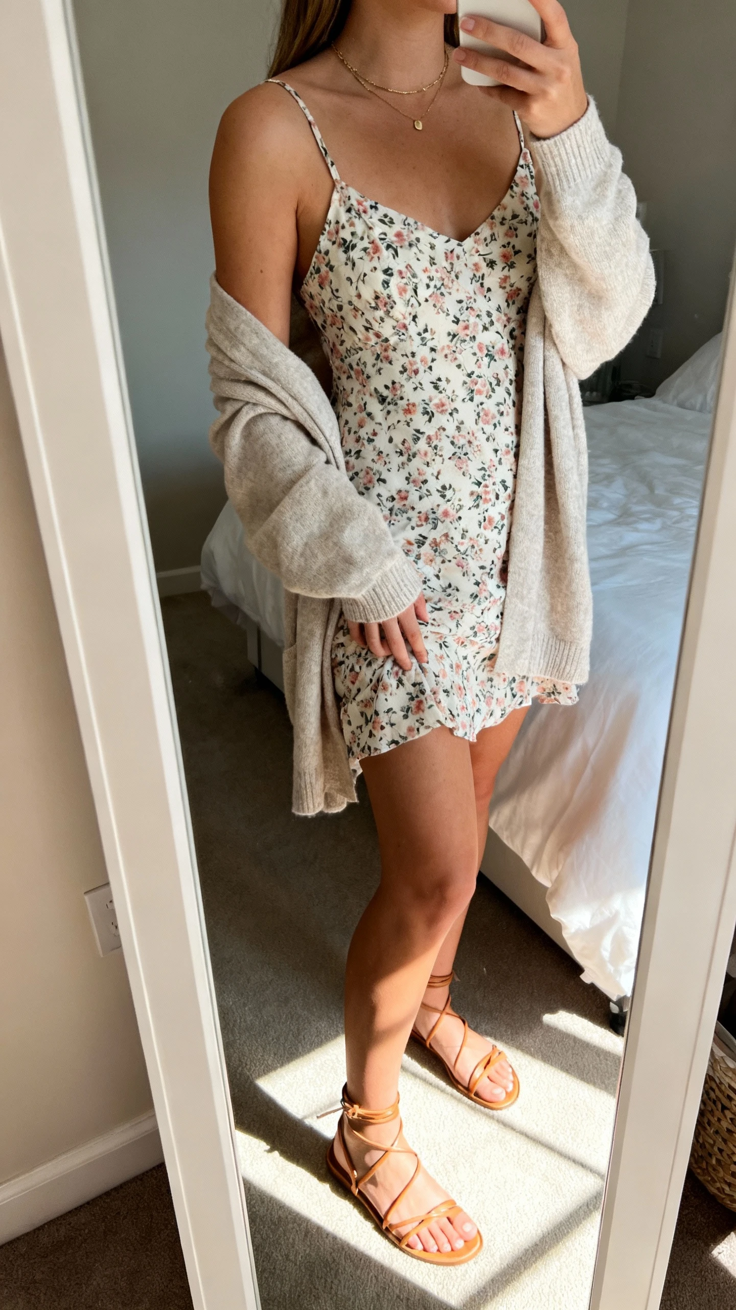 iPhone side-angle mirror selfie of a woman in a floral slip dress with a soft cardigan draped over shoulders, strappy sandals, dainty necklace, face not visible, cropped at shoulders, bedroom mirror with sunny morning light, iPhone photo quality.